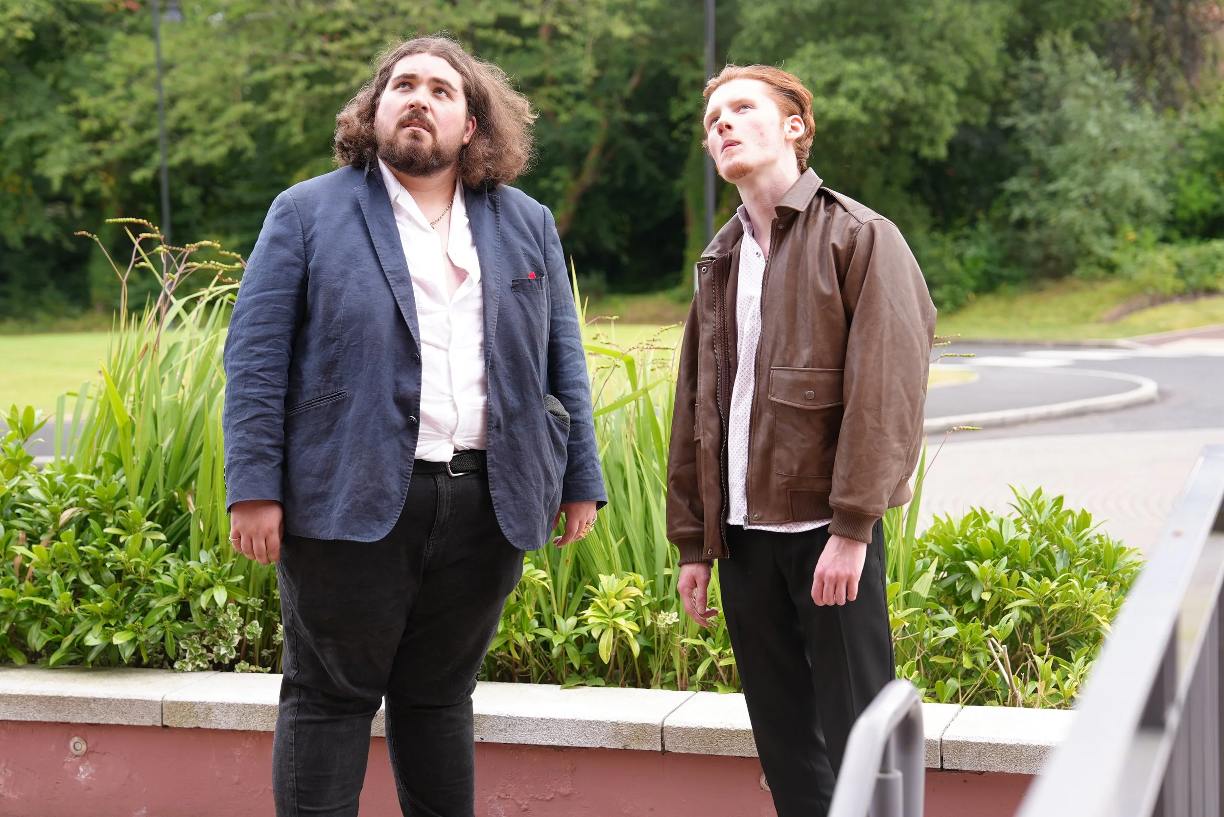 Two young men standing outdoors among greenery, looking up with serious expressions.