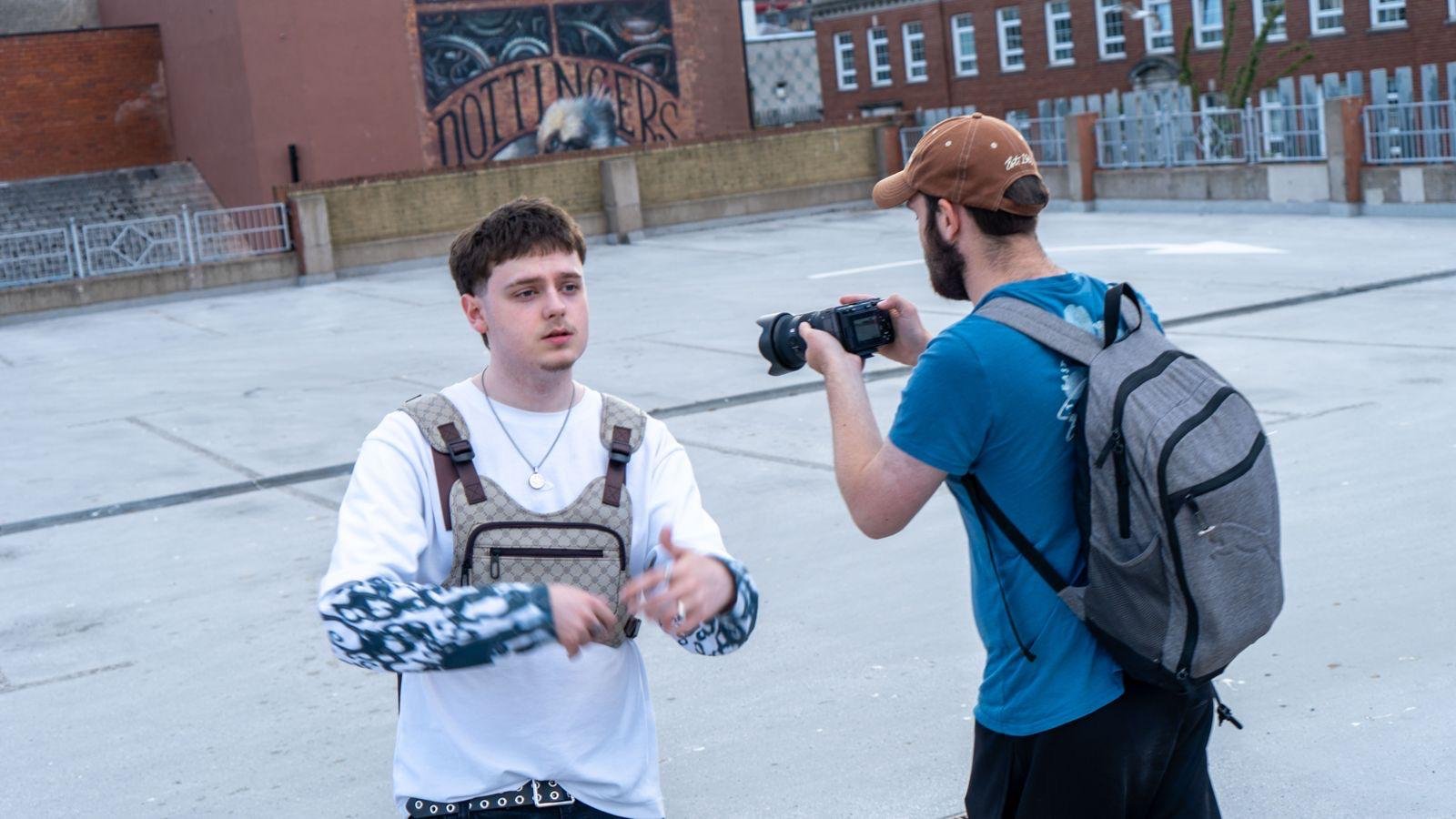 Young man being photographed by a man with a camera in an urban setting where there is a building with graffiti art and a parking lot.