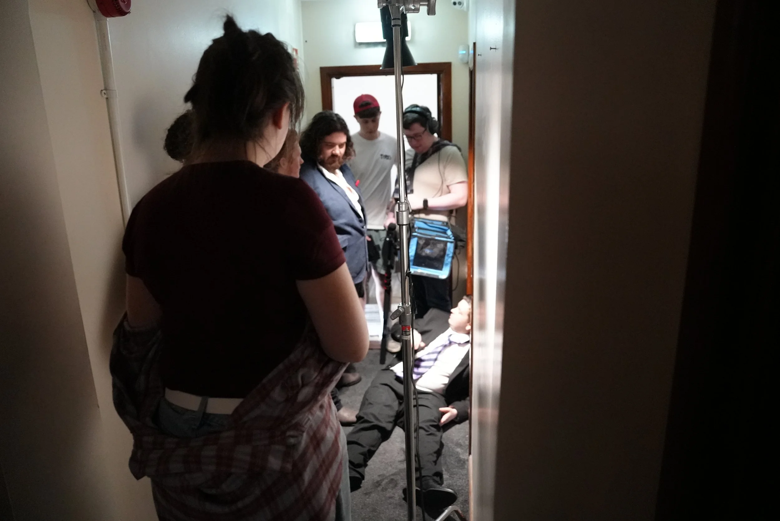 People attending to a person lying on the floor in a narrow hallway, possibly a medical or emergency situation, with three individuals standing and appearing to assist or evaluate.