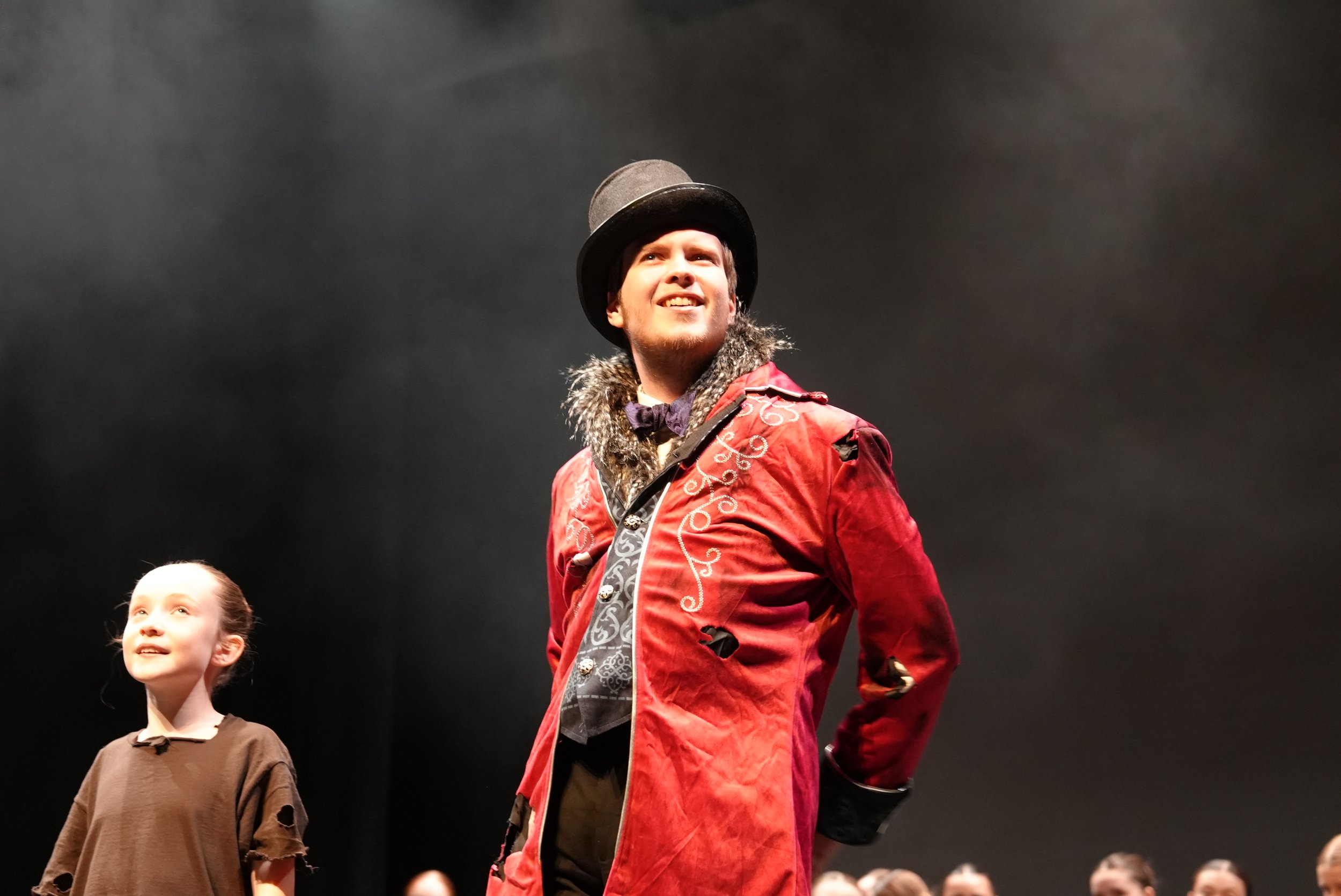 A young man dressed as a circus performer wearing a black top hat, a red velvet jacket with white embroidery, and a patterned vest, smiling and looking upwards. A young girl standing nearby is looking in the same direction, wearing a brown top.