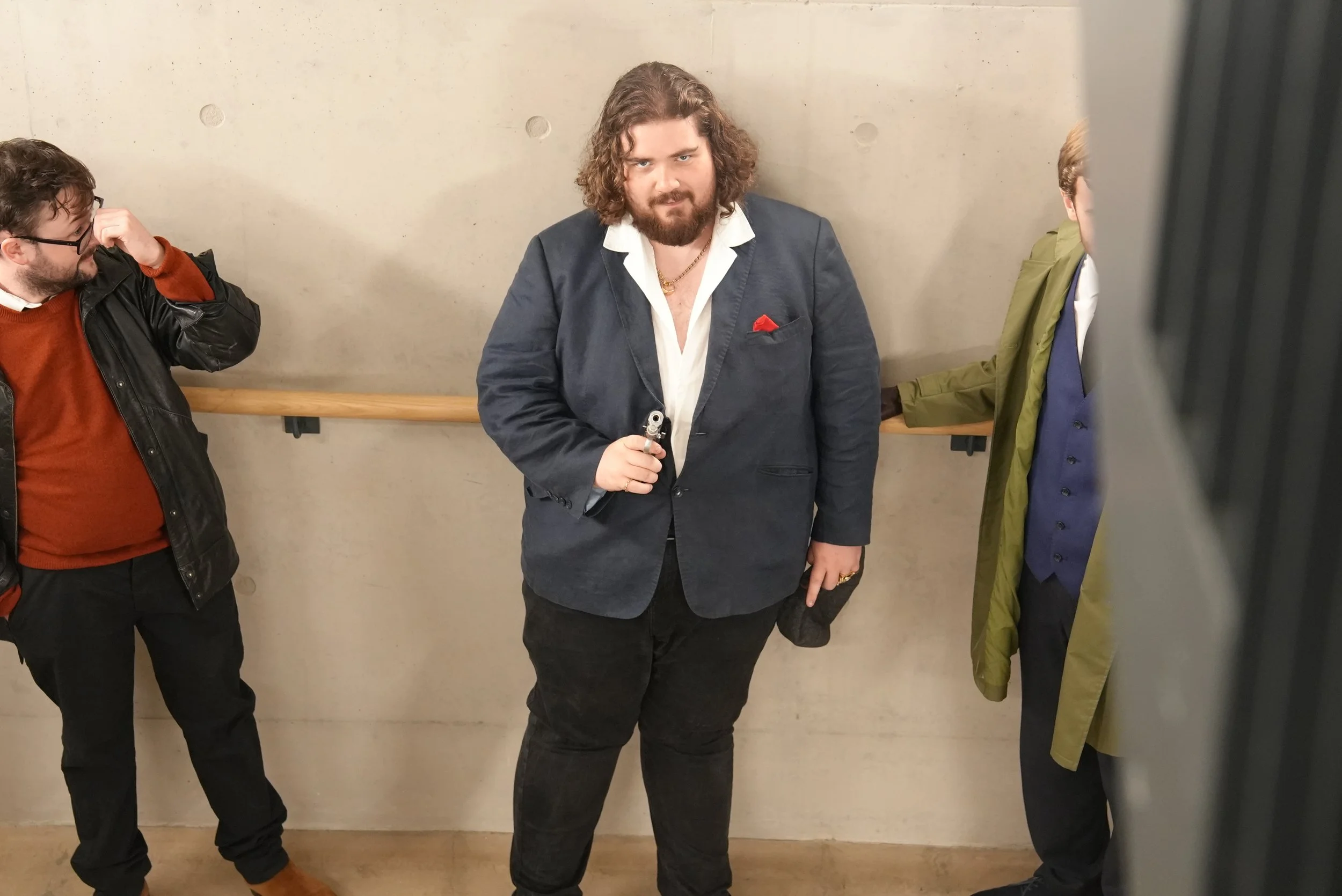 Three men standing against a concrete wall, one in the middle holding a gun and looking directly at the camera, the other two on either side with faces partially visible.