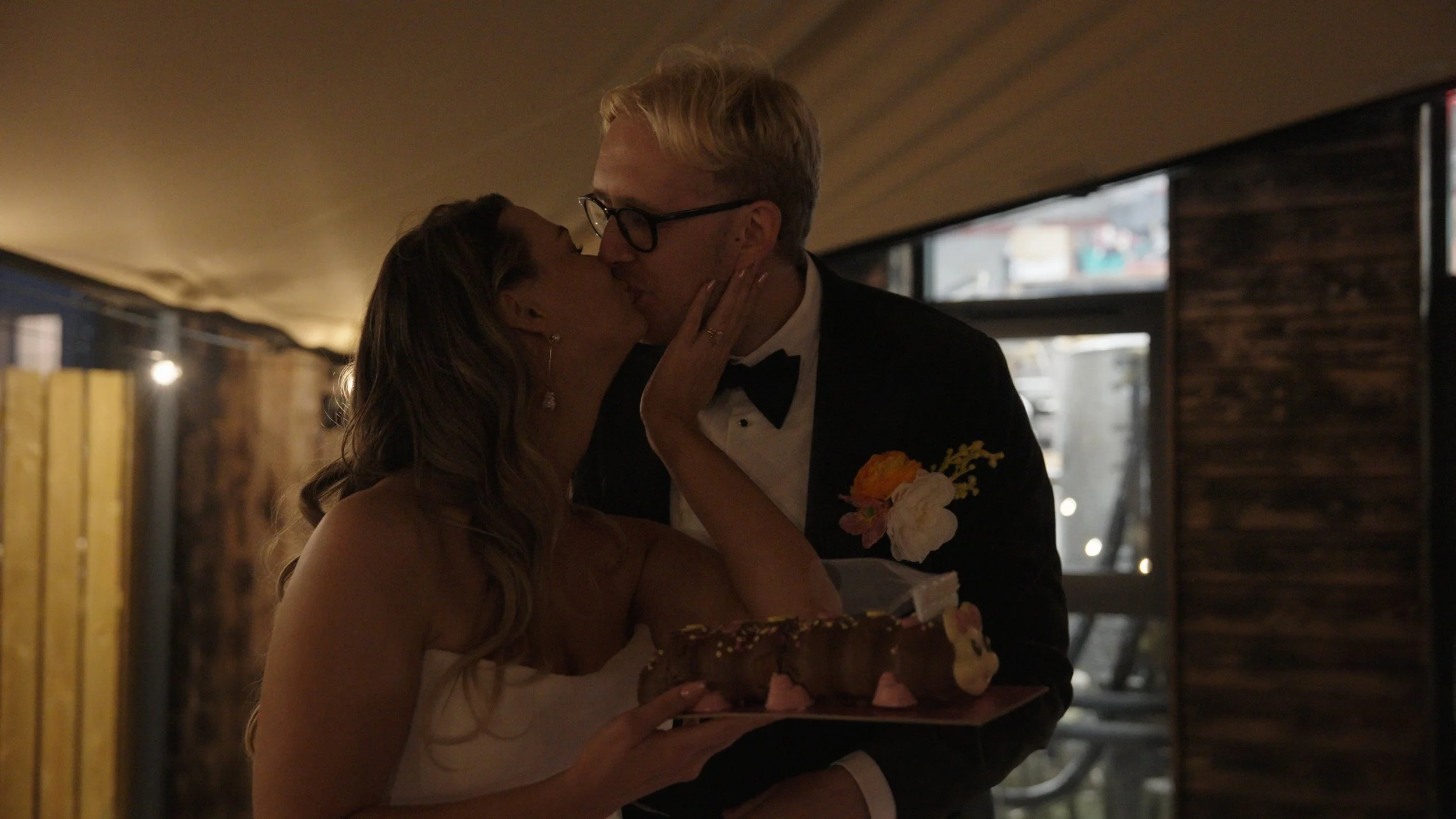 A woman dressed in a wedding gown sharing a kiss with a man dressed in a tuxedo during a wedding celebration, with the woman holding a birthday cake.