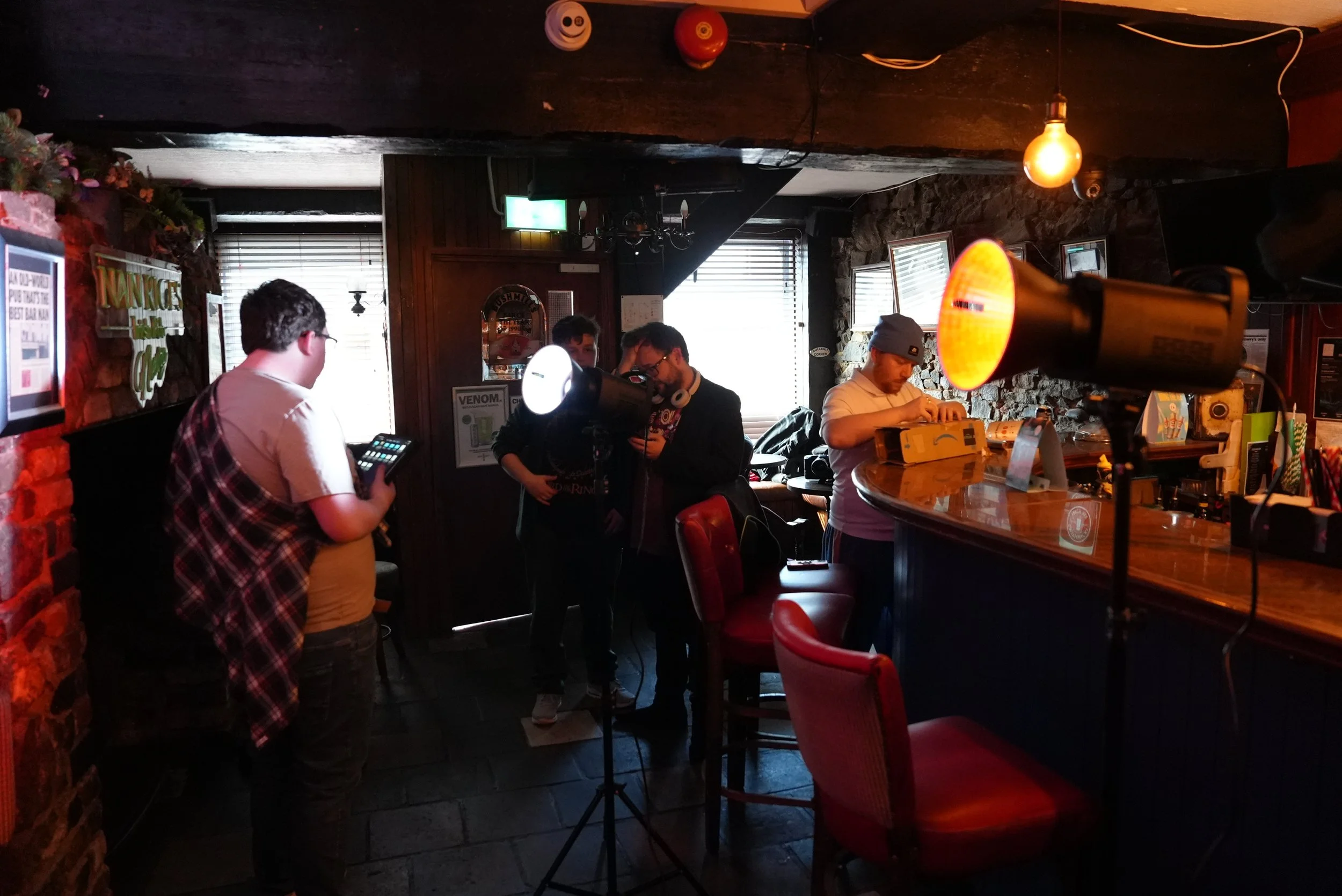 People gathered inside a bar or cafe near the counter, with one person holding a phone and others operating electronic equipment, ambient lighting and rustic interior decor.