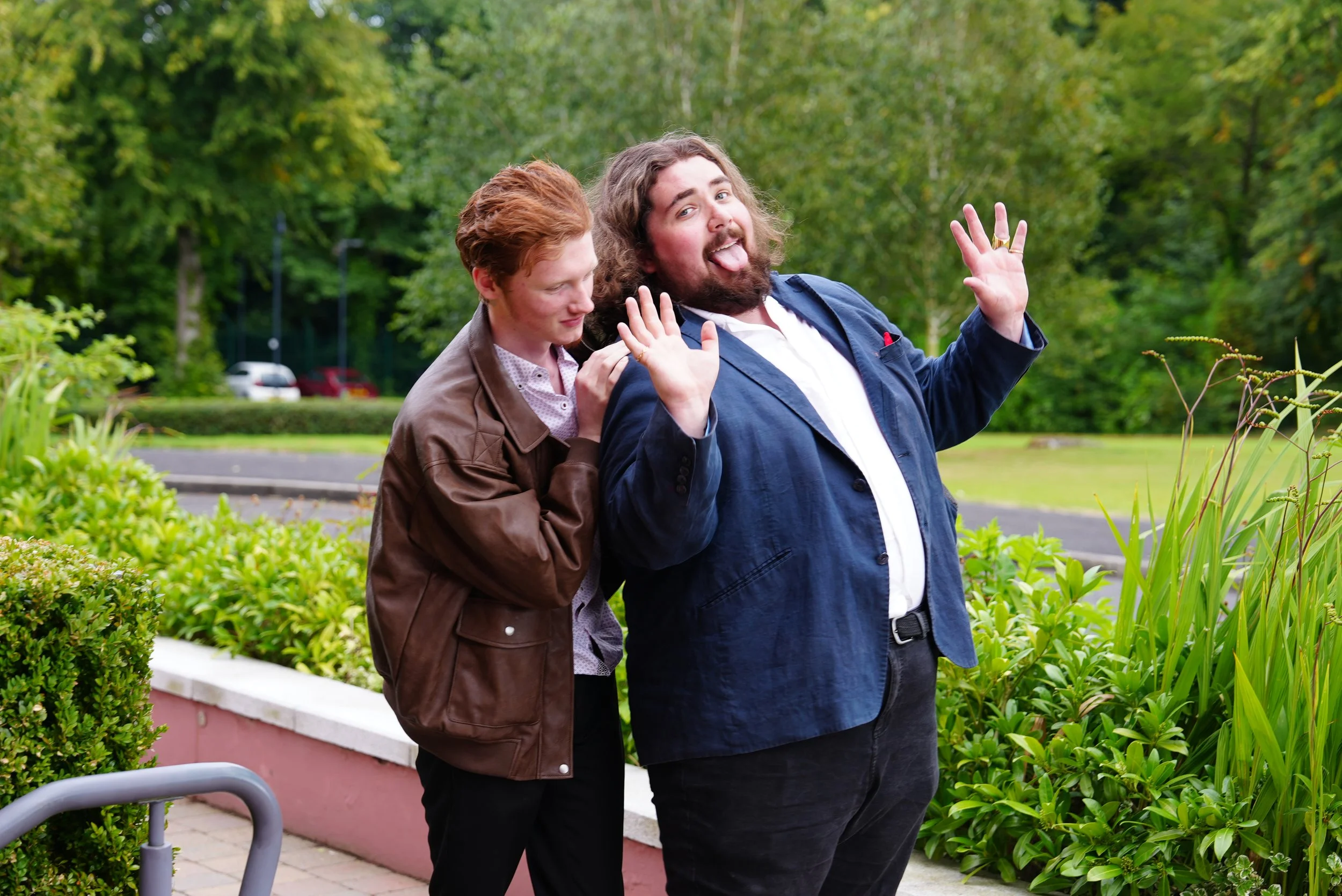 Two men standing outdoors in a green park, with one man having long curly hair and a beard, sticking out his tongue and waving, while the other has short red hair, leaning on his shoulder. They are smiling and making playful gestures.