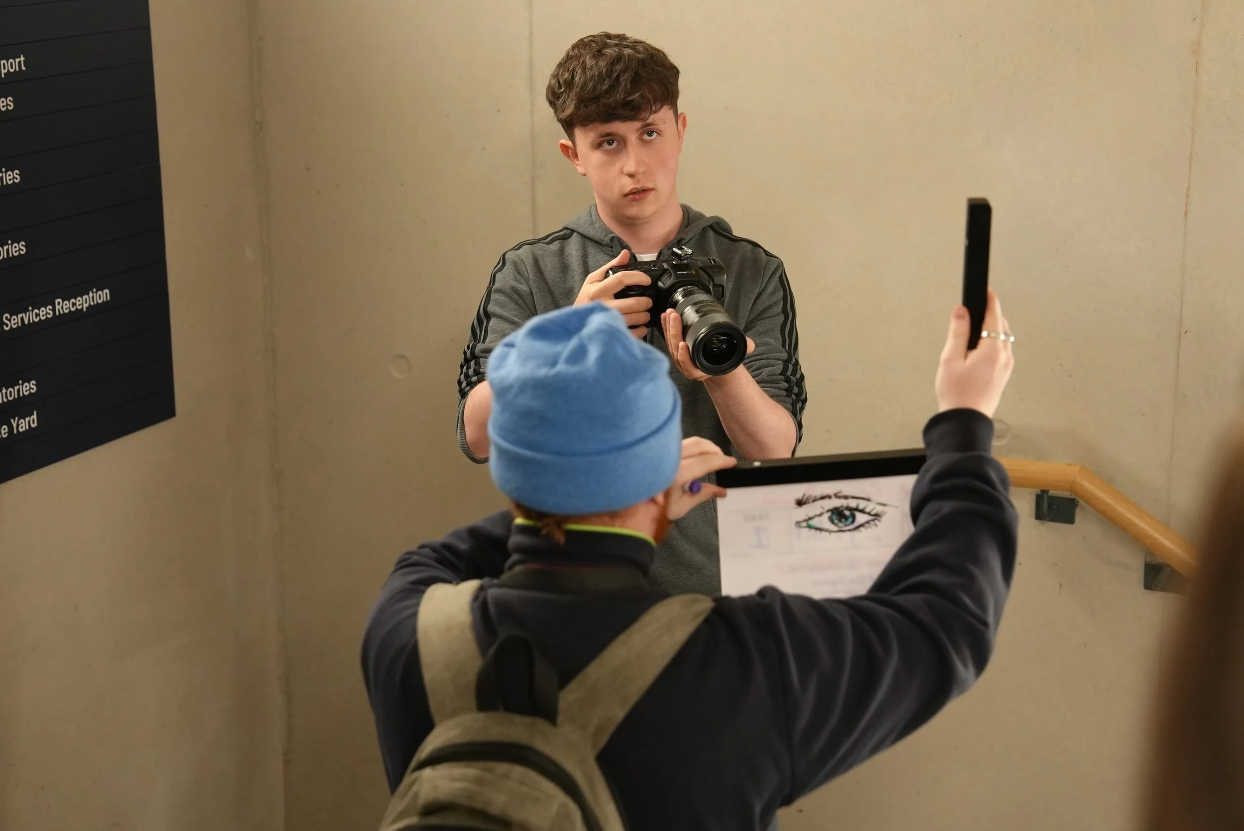 A young man holding a camera looks at a person taking a photo of him, with a person in a blue knit hat and striped shirt holding a tablet with a drawing of an eye on it, taking a photo. The scene appears to be indoors, possibly in a lobby or entrance