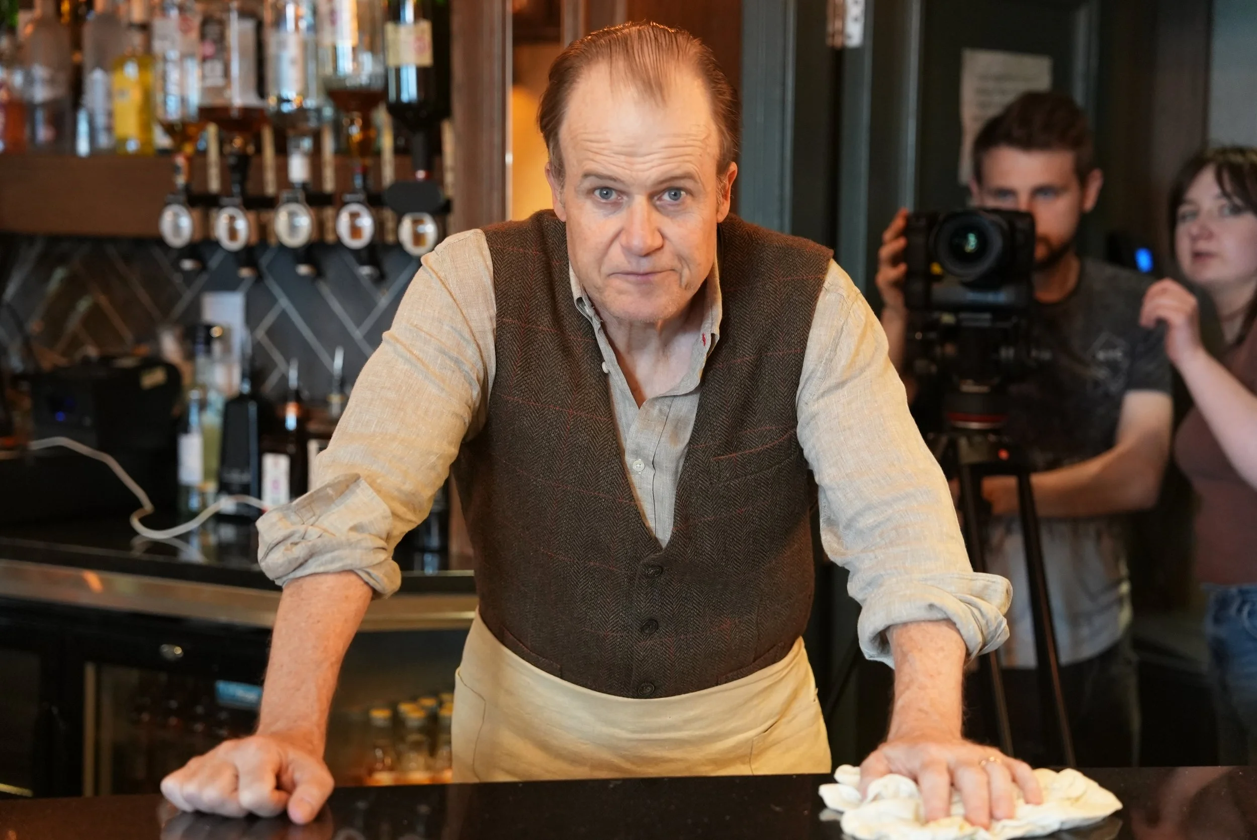 A man with a serious expression leaning forward with his hands on a counter, with two people in the background, one holding a camera, inside a bar or restaurant with bottles on shelves.