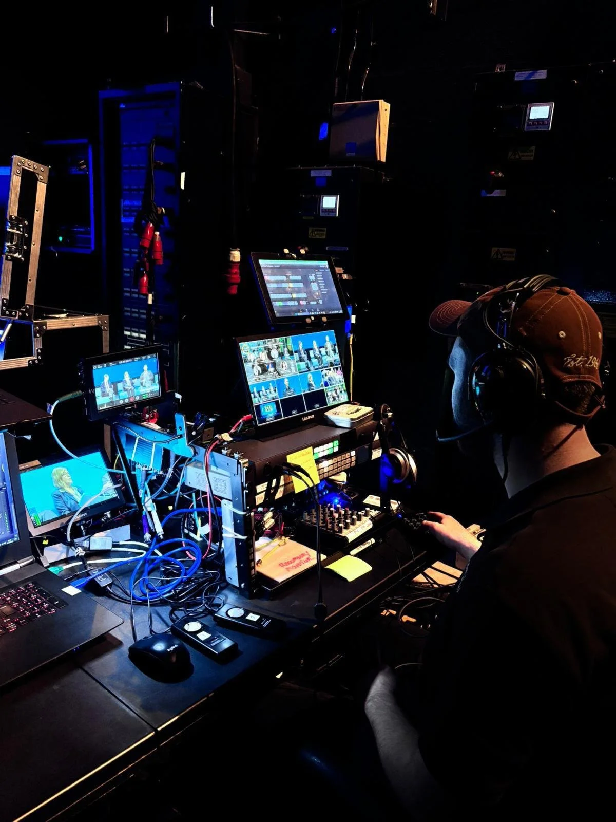 A person operating video production equipment in a dark studio, surrounded by multiple monitors, control panels, and cables.