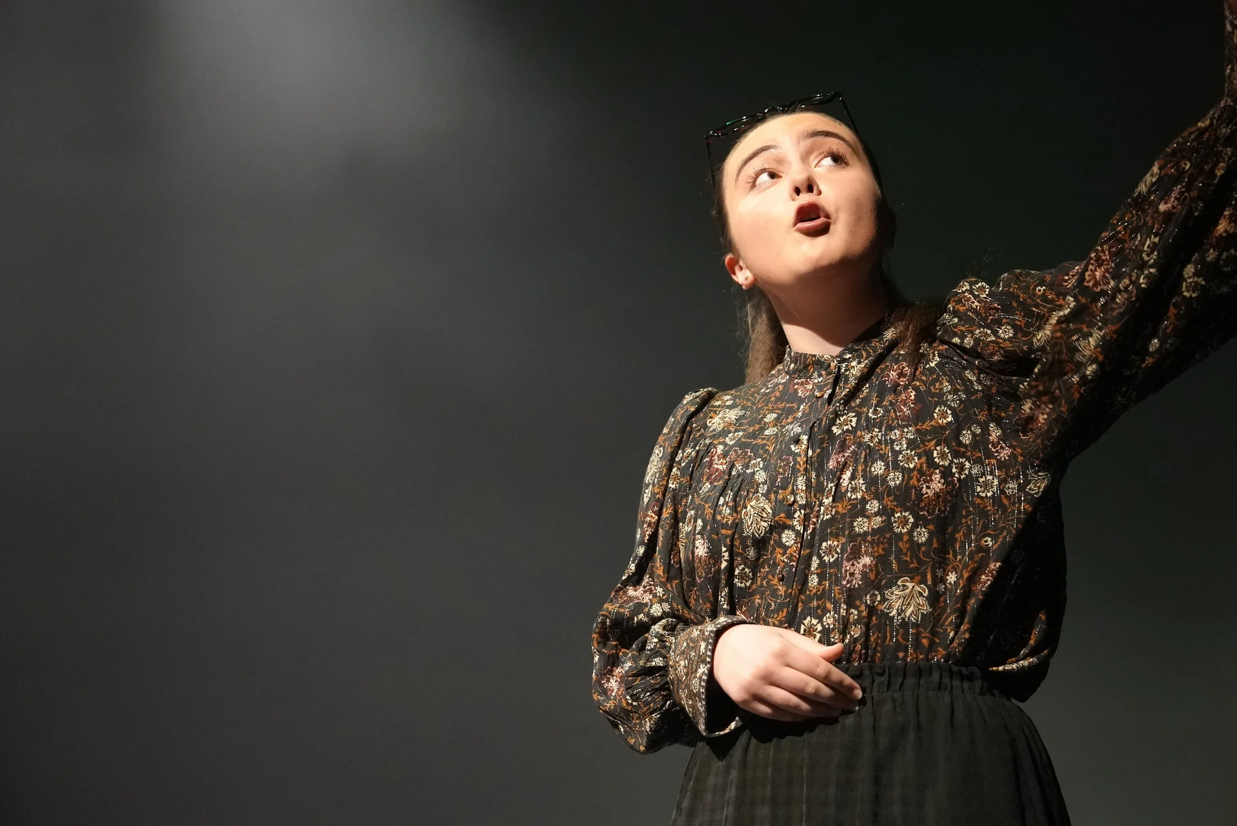 Young woman with glasses on her head wearing a floral patterned blouse and dark skirt, standing against a dark background with one arm raised.