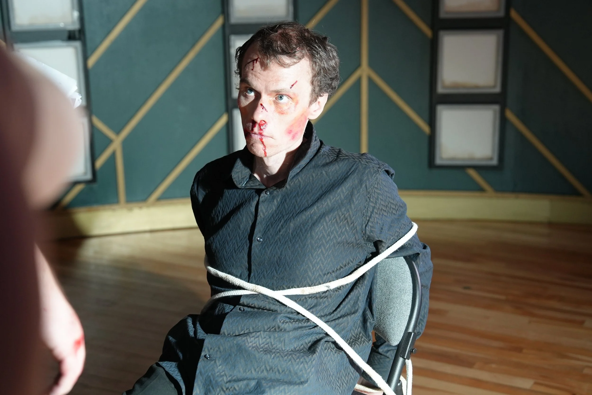 A man with messy hair and bruised, bloodied face tightly bound to a chair with white rope, staring with wide eyes. The background features geometric patterned walls with framed artwork.