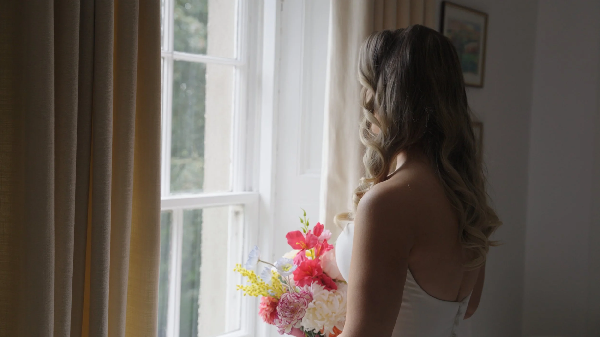 A woman with long wavy hair holding a bouquet of pink and yellow flowers while looking out a window in a softly lit room.