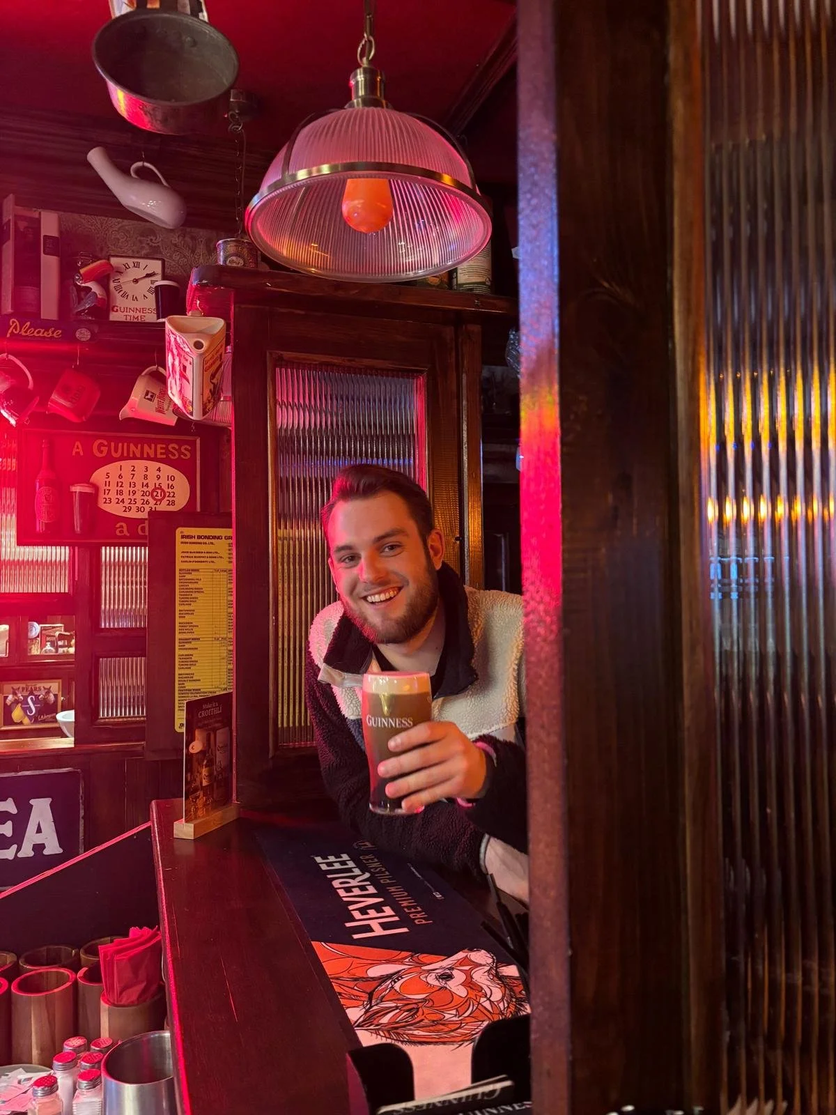A smiling man with a beard holding a pint of Guinness beer in a bar with wood paneling and various Guinness memorabilia on the wall.