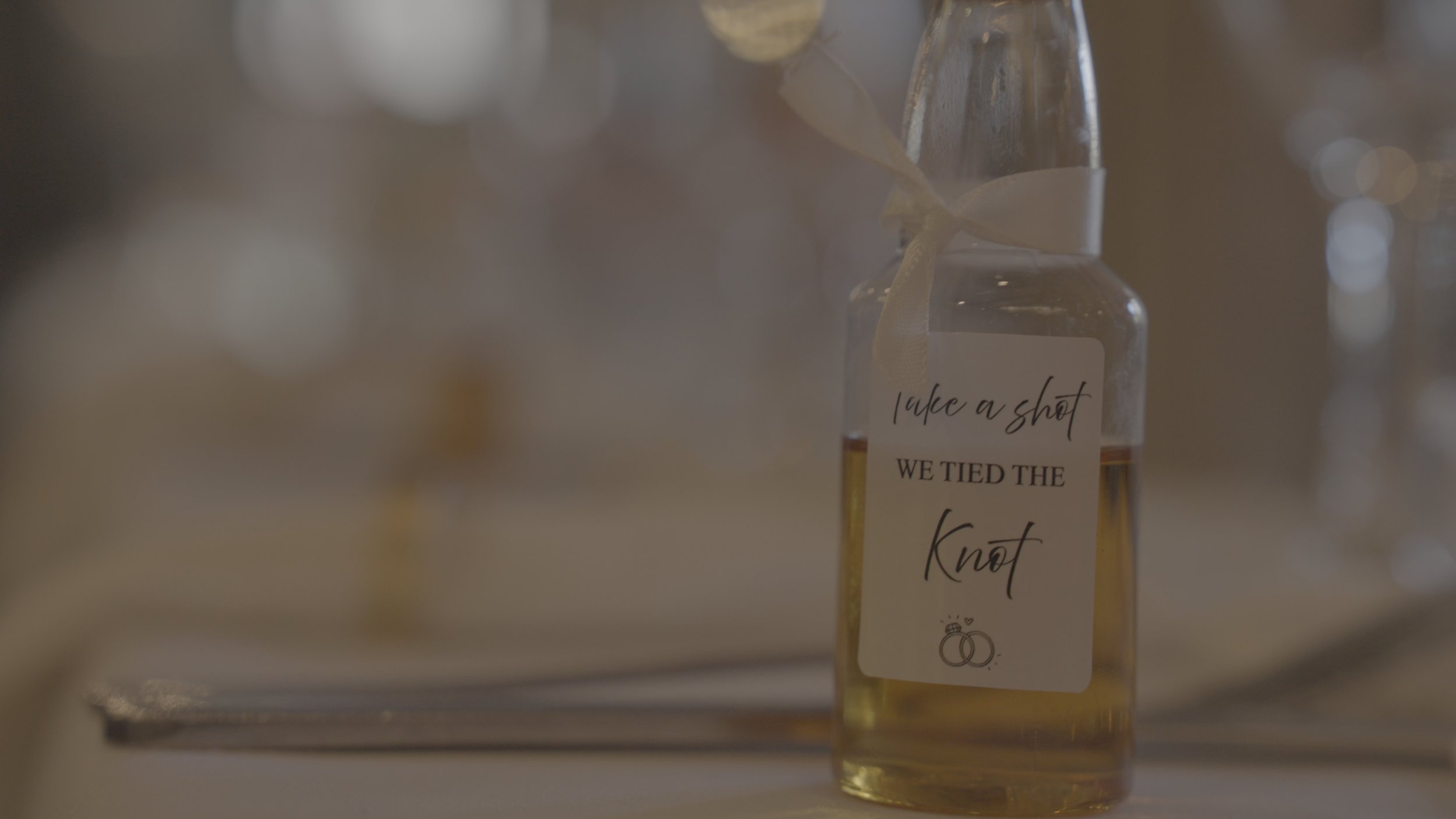 A small glass bottle with a label that reads 'take a shot, we tied the knot', containing a yellow beverage, decorated with a ribbon, surrounded by blurred bottles in the background.