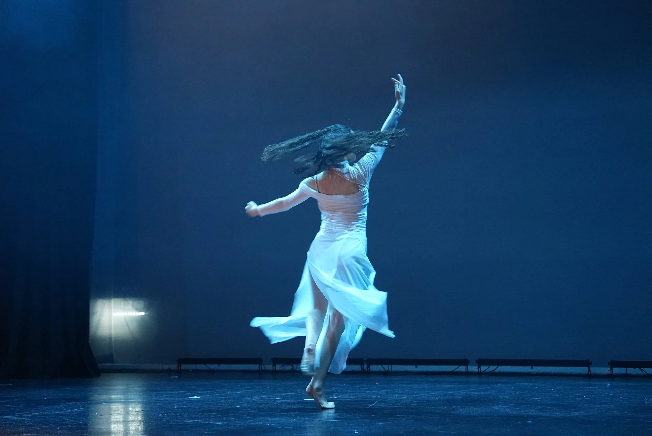 A dancer in a white flowing dress performing a dance on stage with a dark background.