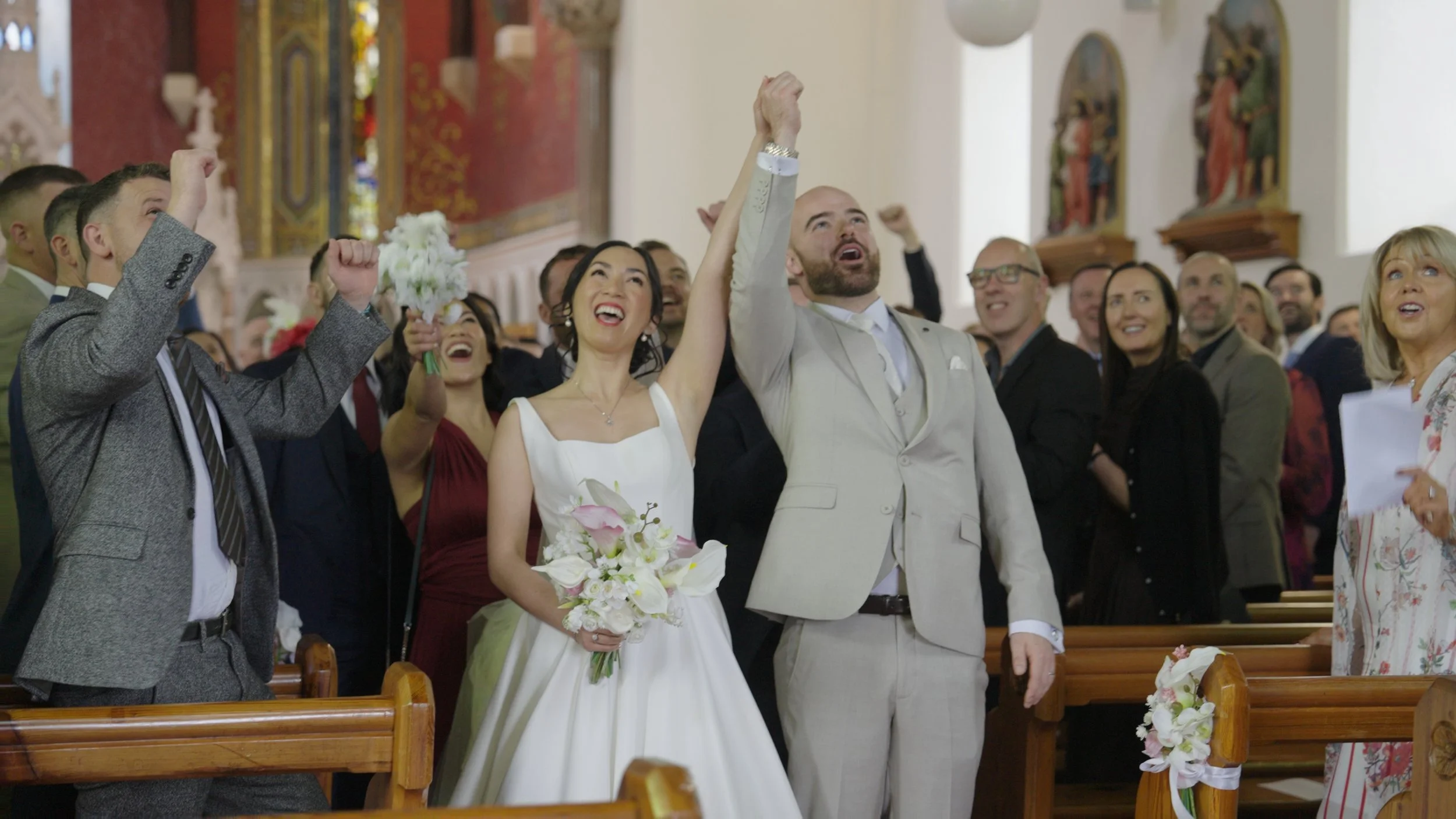 A bride and groom raising their fists in celebration inside a church, surrounded by smiling guests.