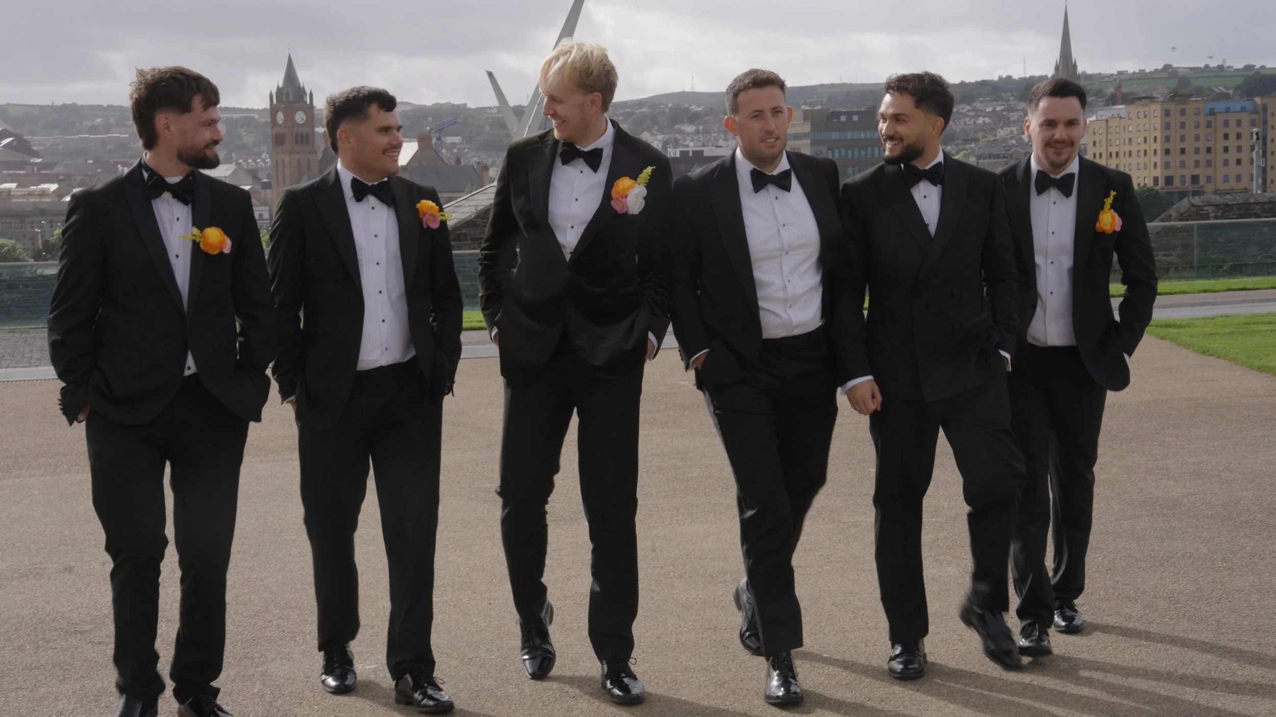 Six men in tuxedos with boutonnières, walking together outdoors on a cloudy day against a cityscape with church steeples and modern buildings.