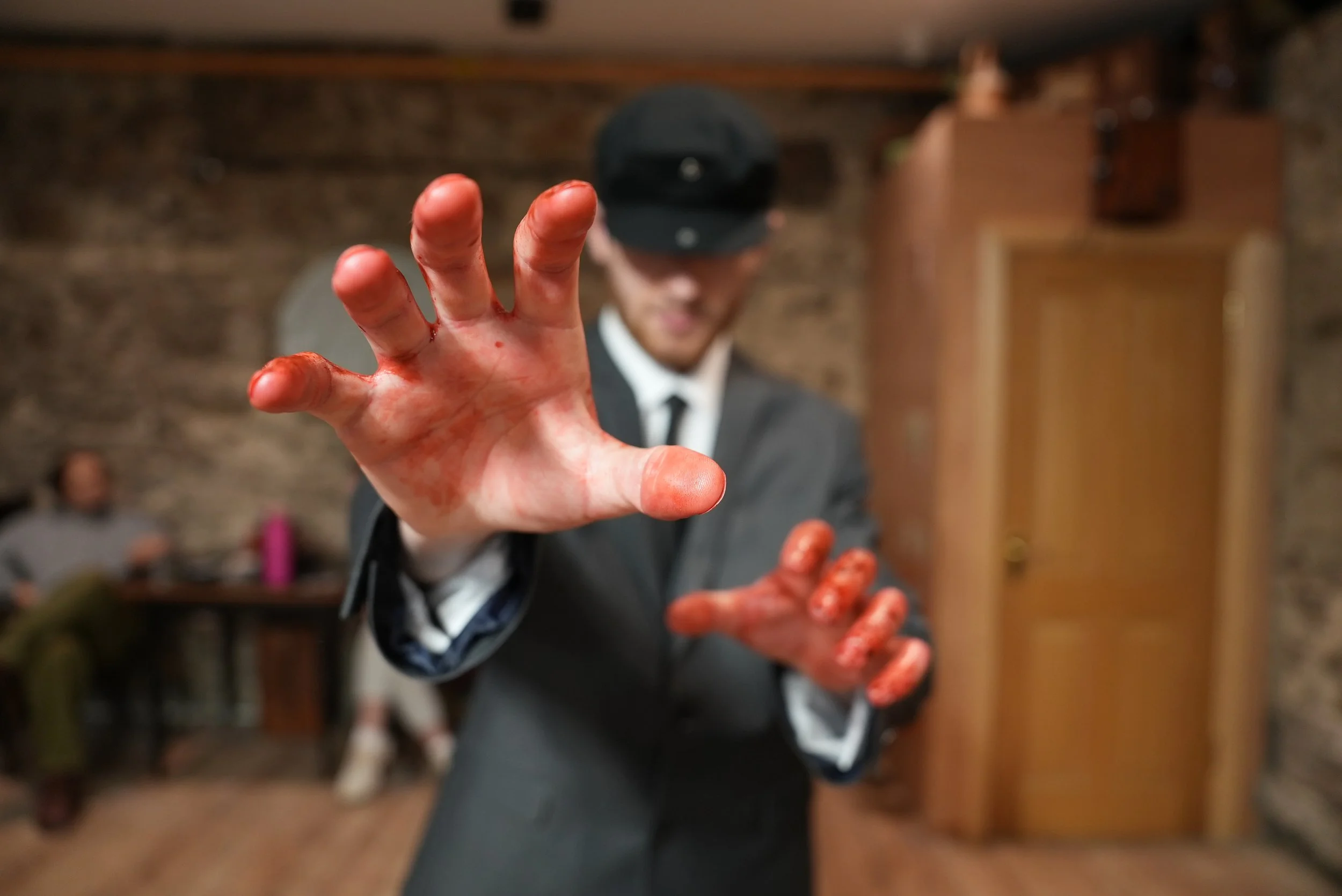 A person wearing a suit and a cap reaching out with a blood-stained hand toward the camera in a cozy, wood-paneled room with blurred background.