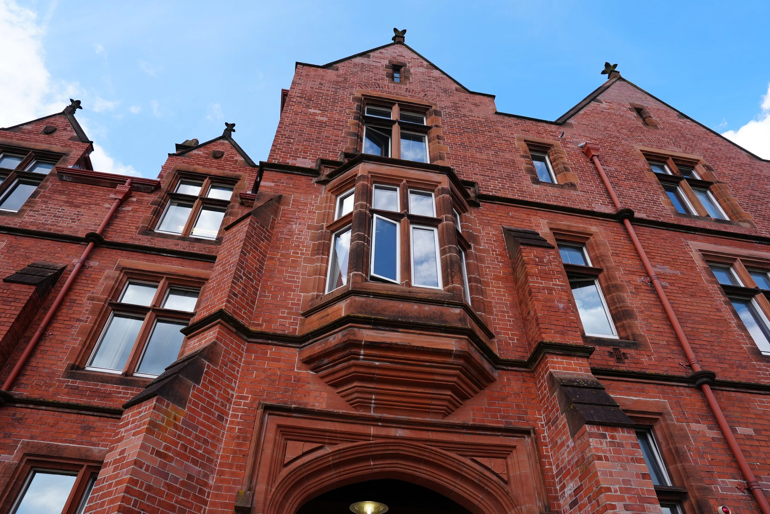 A low-angle view of a red brick gothic-style building with multiple windows and decorative stone details against a partly cloudy sky.