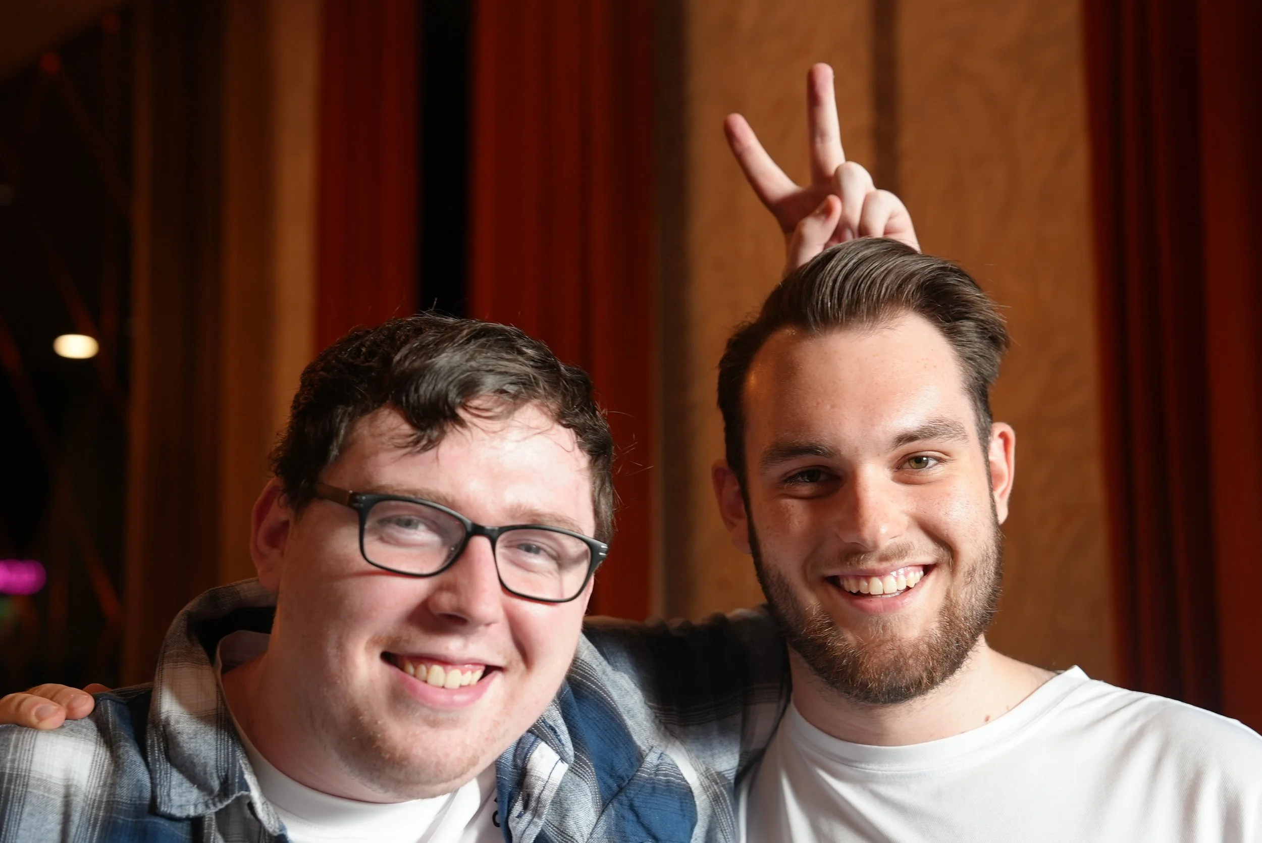 Two men smiling, one with glasses and a plaid shirt, the other with a beard and white shirt, close together in a room with wooden walls.