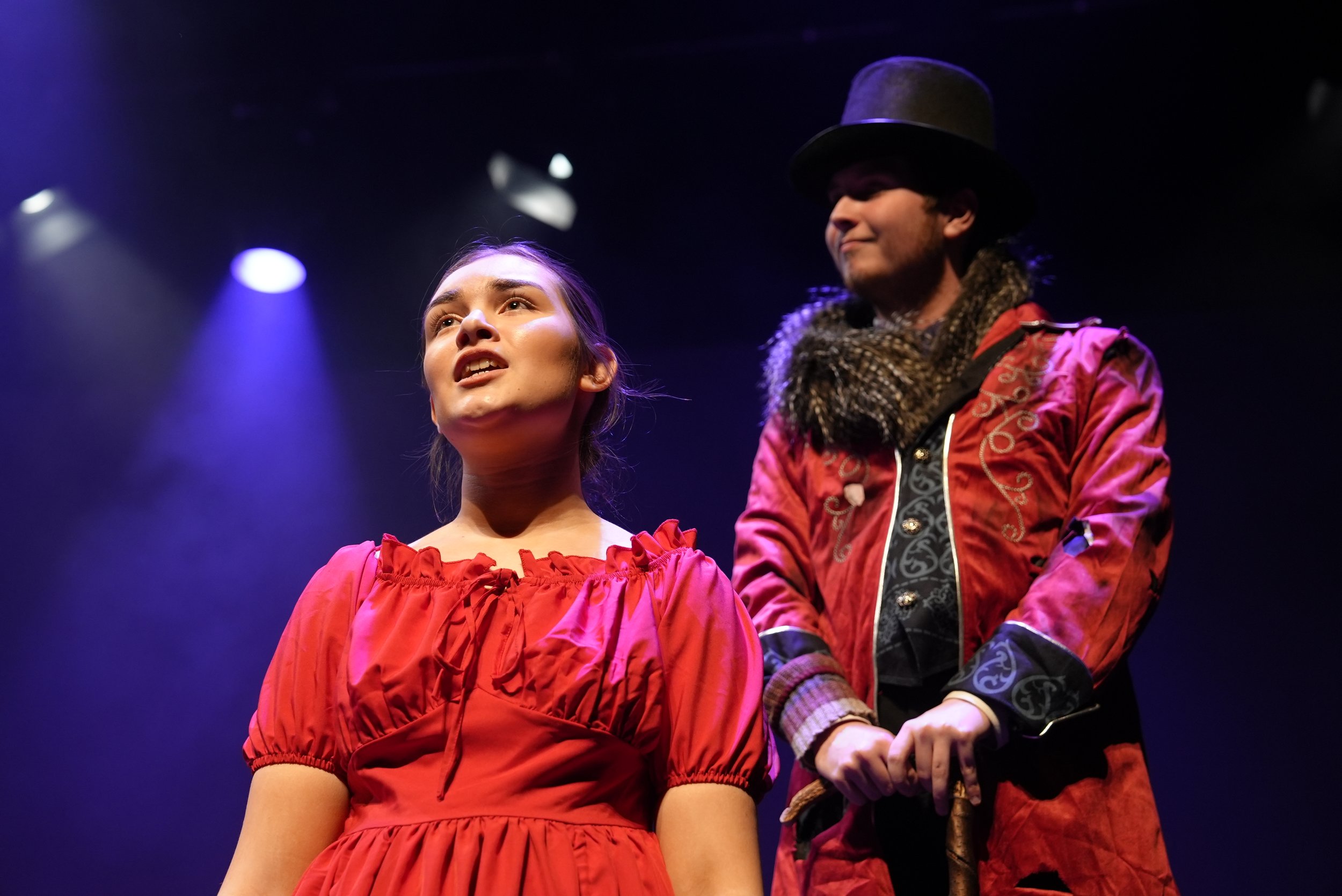 A young woman with light brown hair and a young man with a beard, wearing a top hat and ornate jacket, standing on stage under purple spotlights.