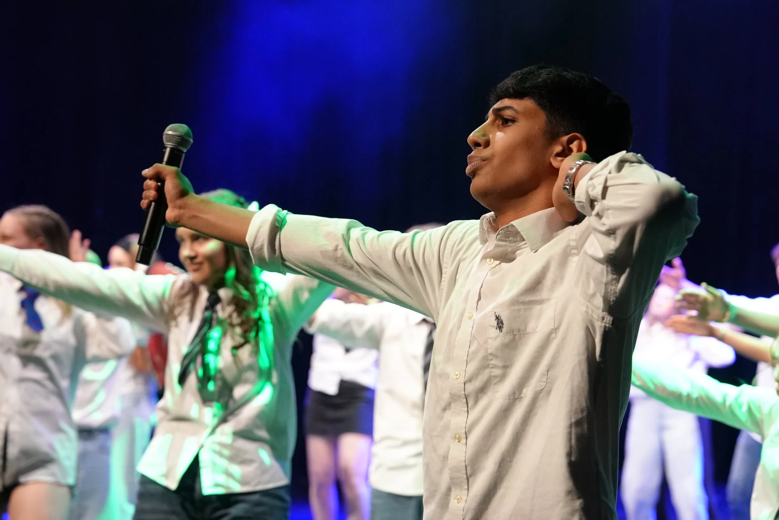 A young man with short black hair, wearing a white button-up shirt, is performing a dance or gesture on stage. He has his arms extended and hands pointed outward, with a serious expression. Behind him, there are other young people participating in th