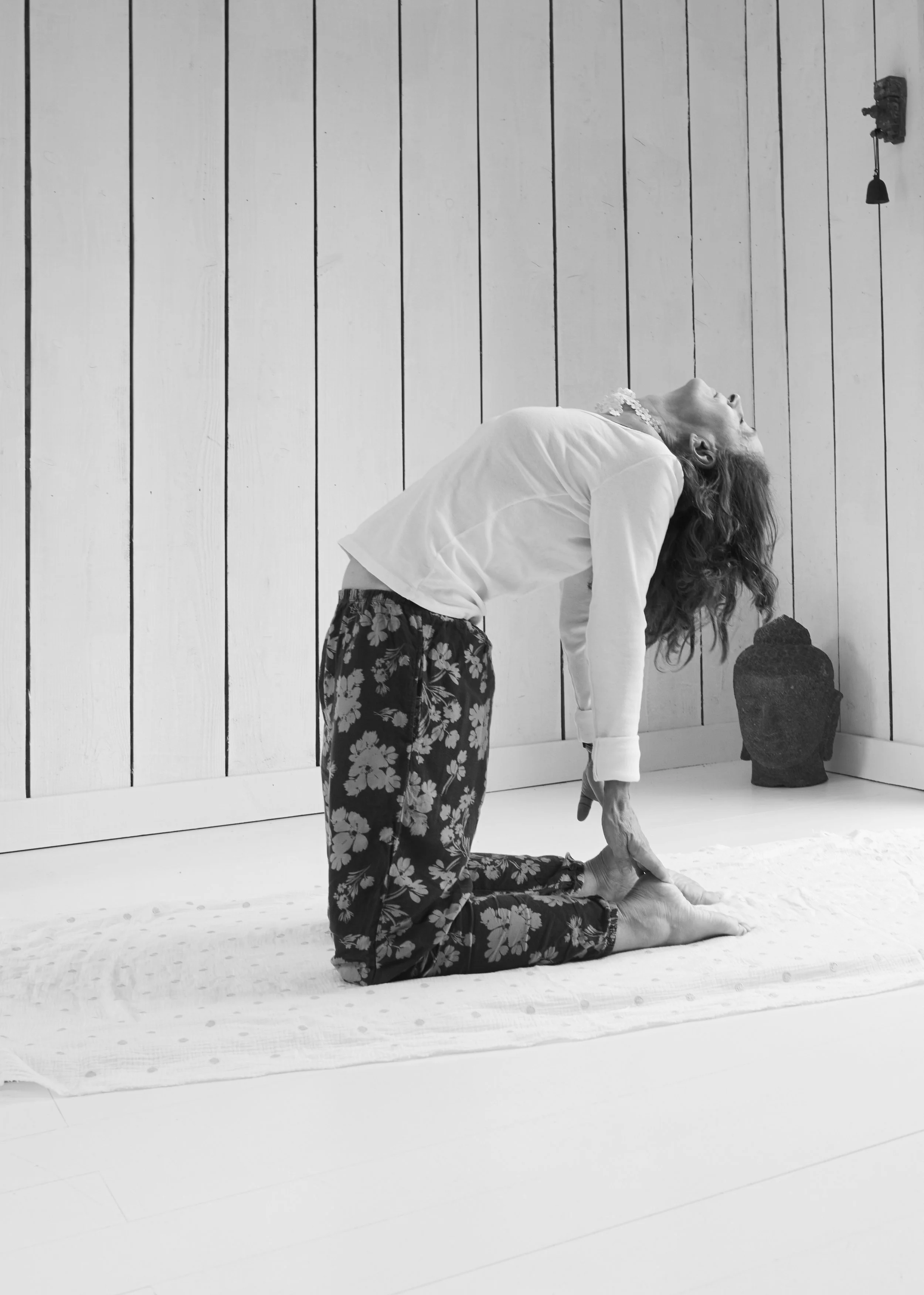 Une femme âgée en position de yoga, à genoux avec le corps penché en avant, dans une pièce avec un mur en bois clair. Elle porte un haut blanc et un pantalon à motifs floraux noirs et blancs. Il y a un tapis de yoga et un grand vase en potière à côté d'elle.