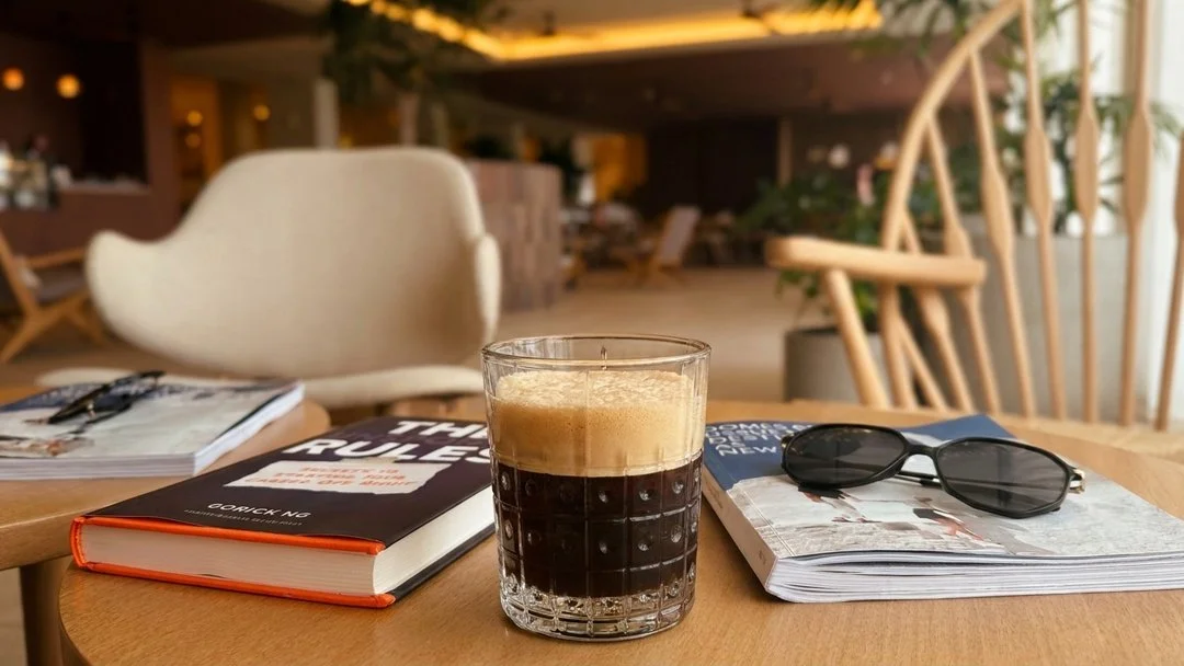 A glass of dark beer with foam on top placed on a wooden table. Next to the glass are two books, one titled 'The Rule' and the other with sunglasses resting on it. The background shows a cozy indoor seating area with chairs and plants.