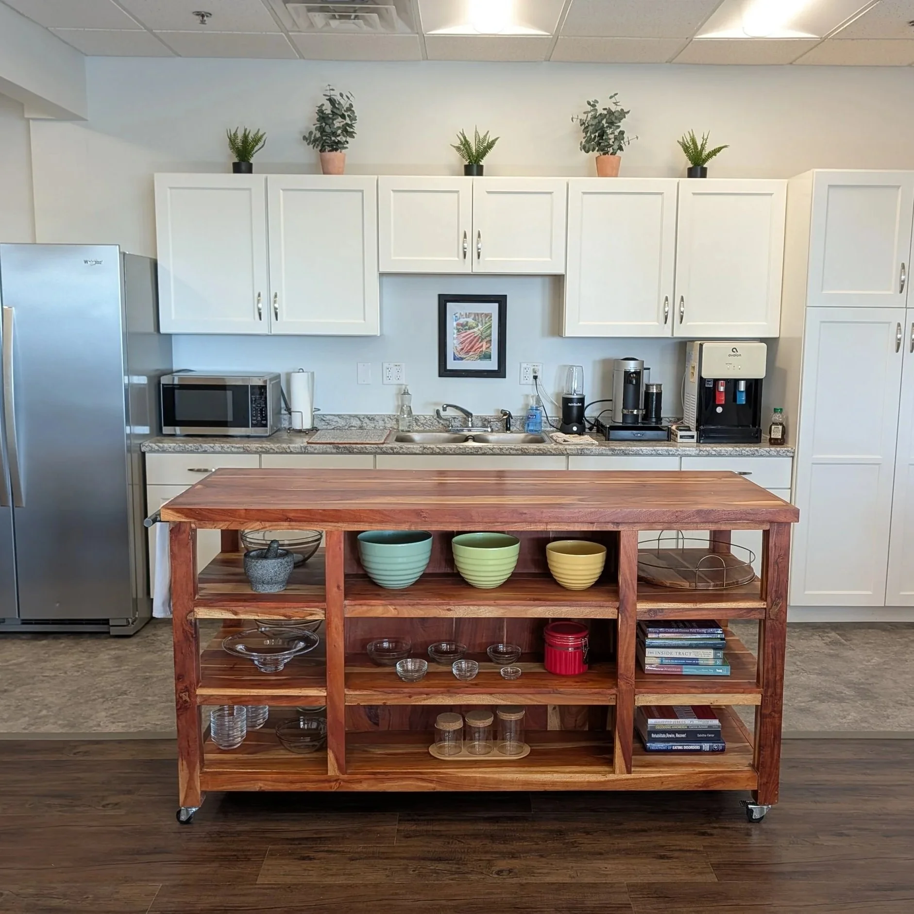 A kitchen with white cabinets, a granite countertop, and a wooden island on wheels with shelves holding glassware and bowls. There is a microwave, coffee maker, water dispenser, and small framed art on the wall.