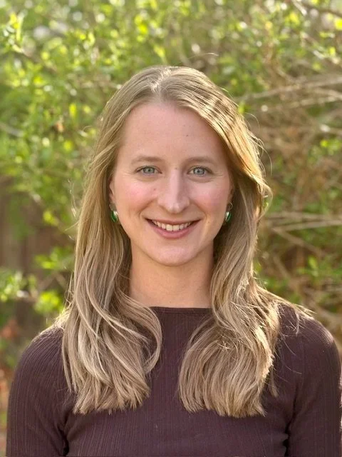 A woman with blonde hair and green earrings smiling outdoors with trees and greenery in the background.