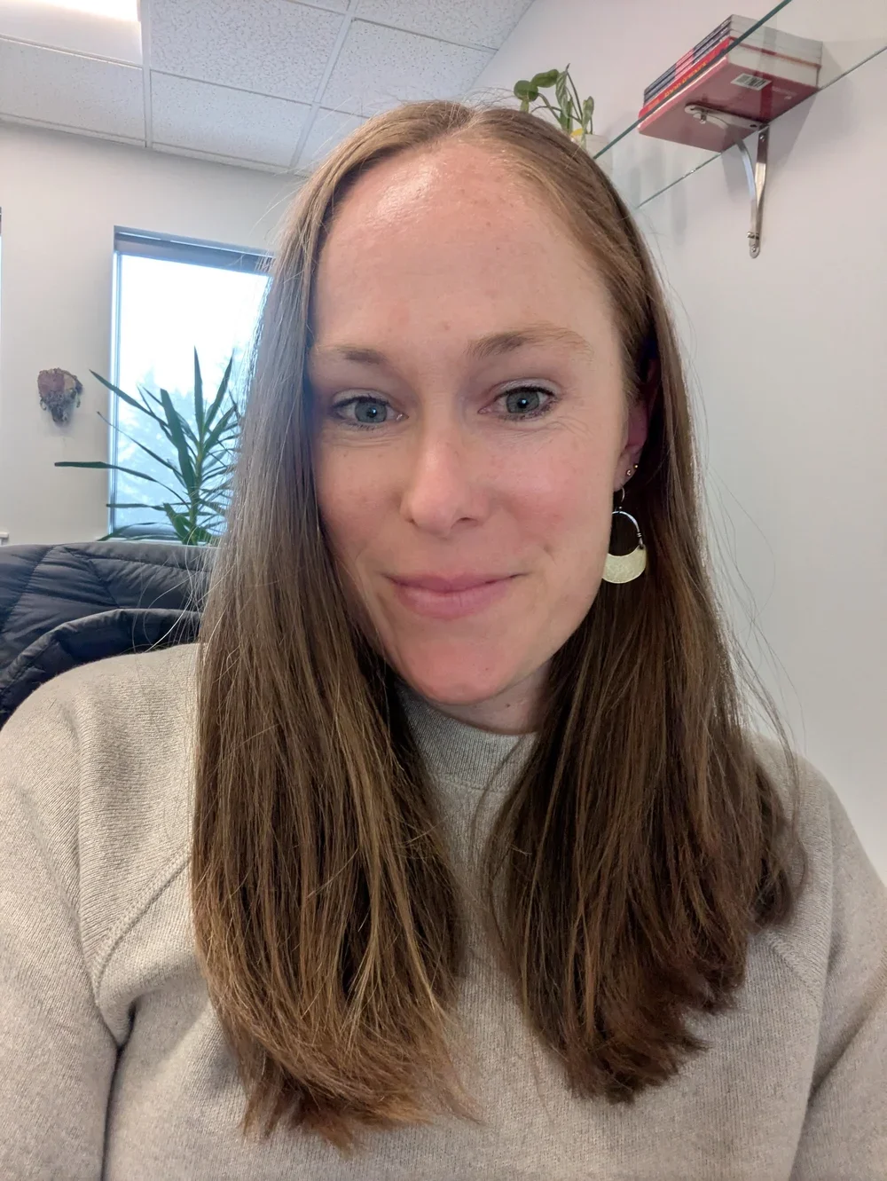 A woman with long brown hair and earrings, smiling in an office setting with a potted plant, window, and wall shelf in the background.