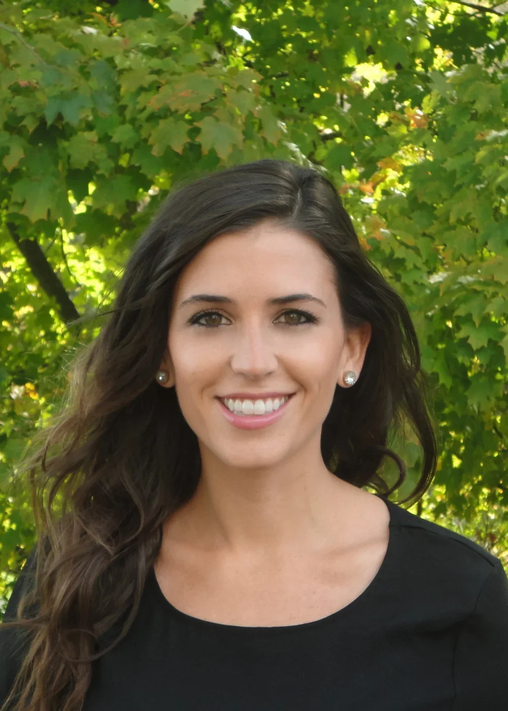 A young woman with long, wavy dark hair smiling outdoors, wearing a black top and pearl earrings, with green trees in the background.