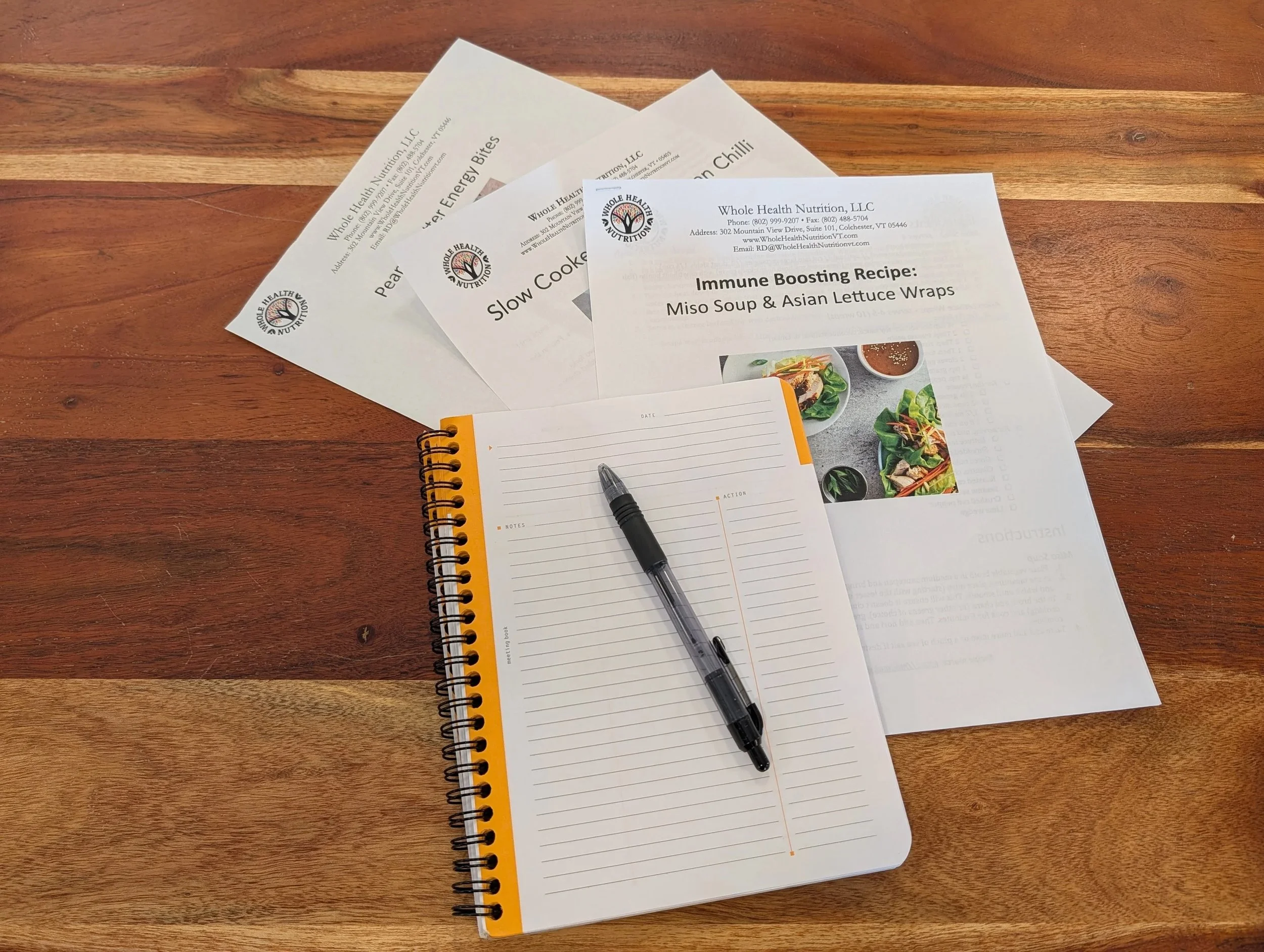Three sheets of paper with the Whole Health Nutrition logo, a small spiral notebook, and a pen on a wooden table.