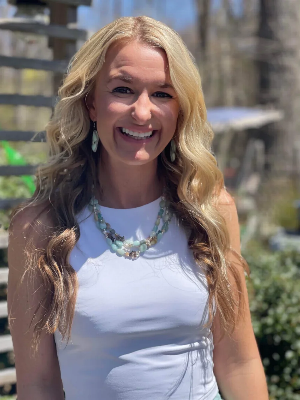 A woman with long, wavy blonde hair smiling outdoors, wearing a white sleeveless top and a necklace with multicolored beads.