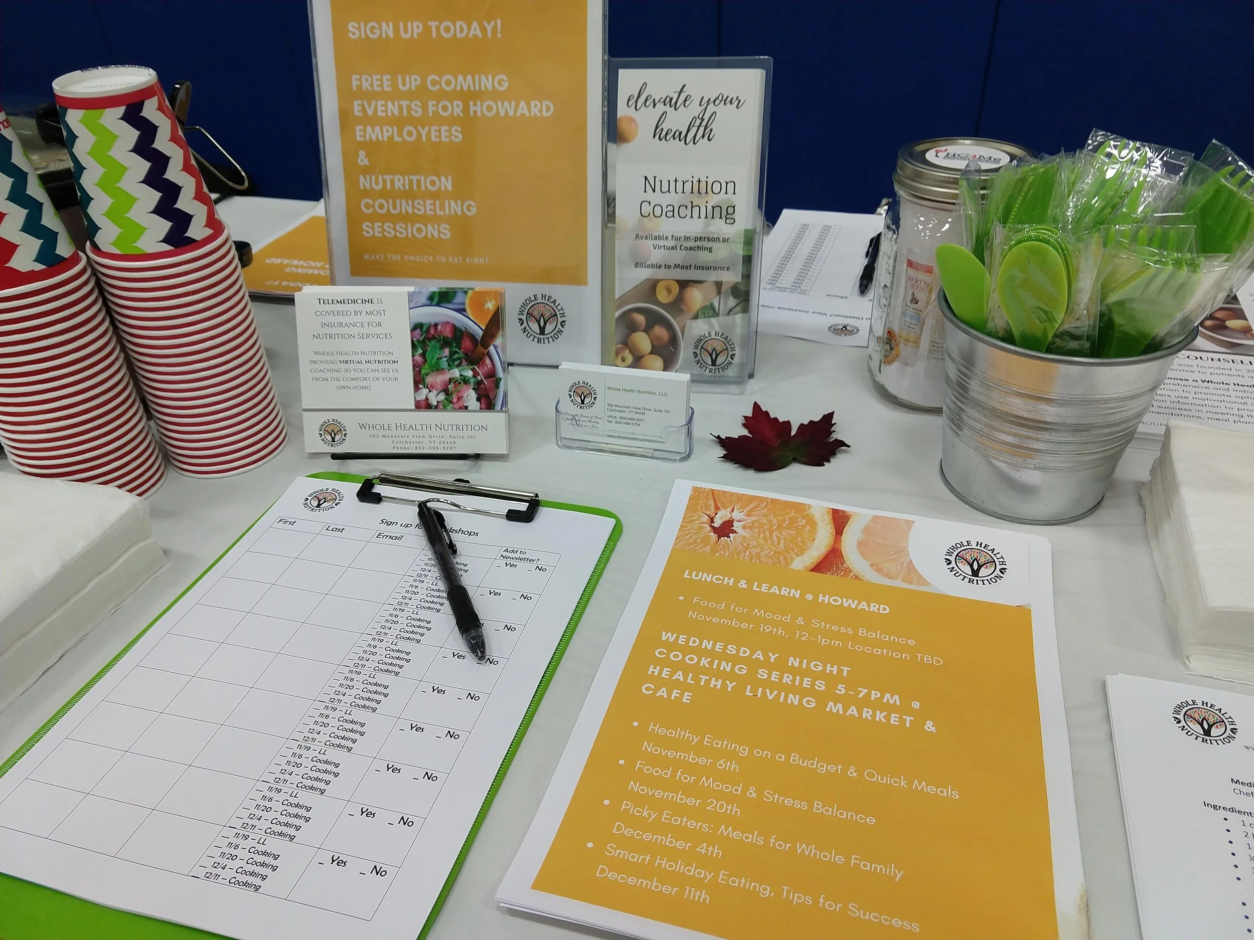 Table with cups, informational signs about health and nutrition coaching, a sign-up sheet, a pen, and green utensils in a container.