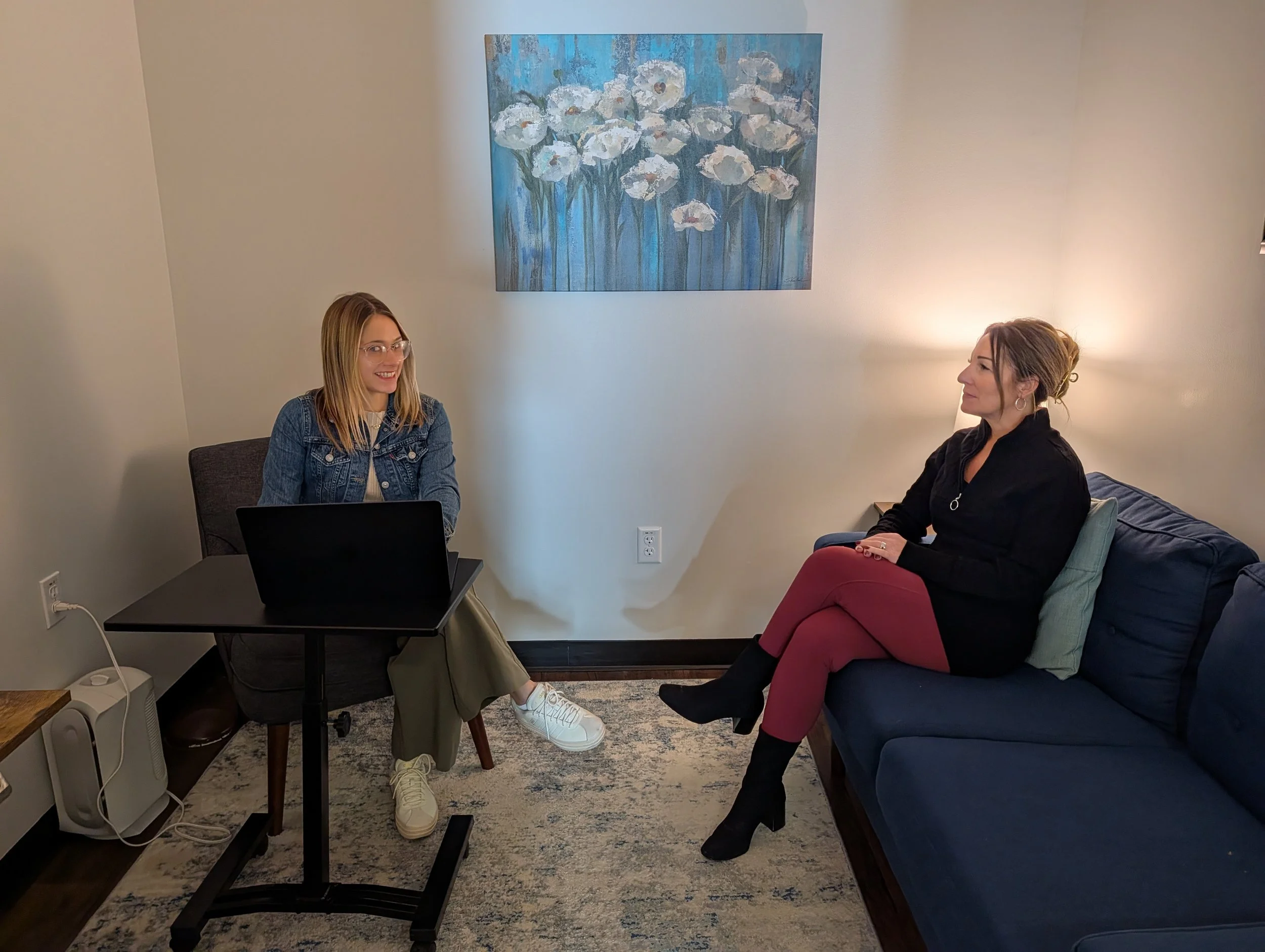Two women sitting and talking in a living room with a painting of white flowers on a blue background hanging on the wall.