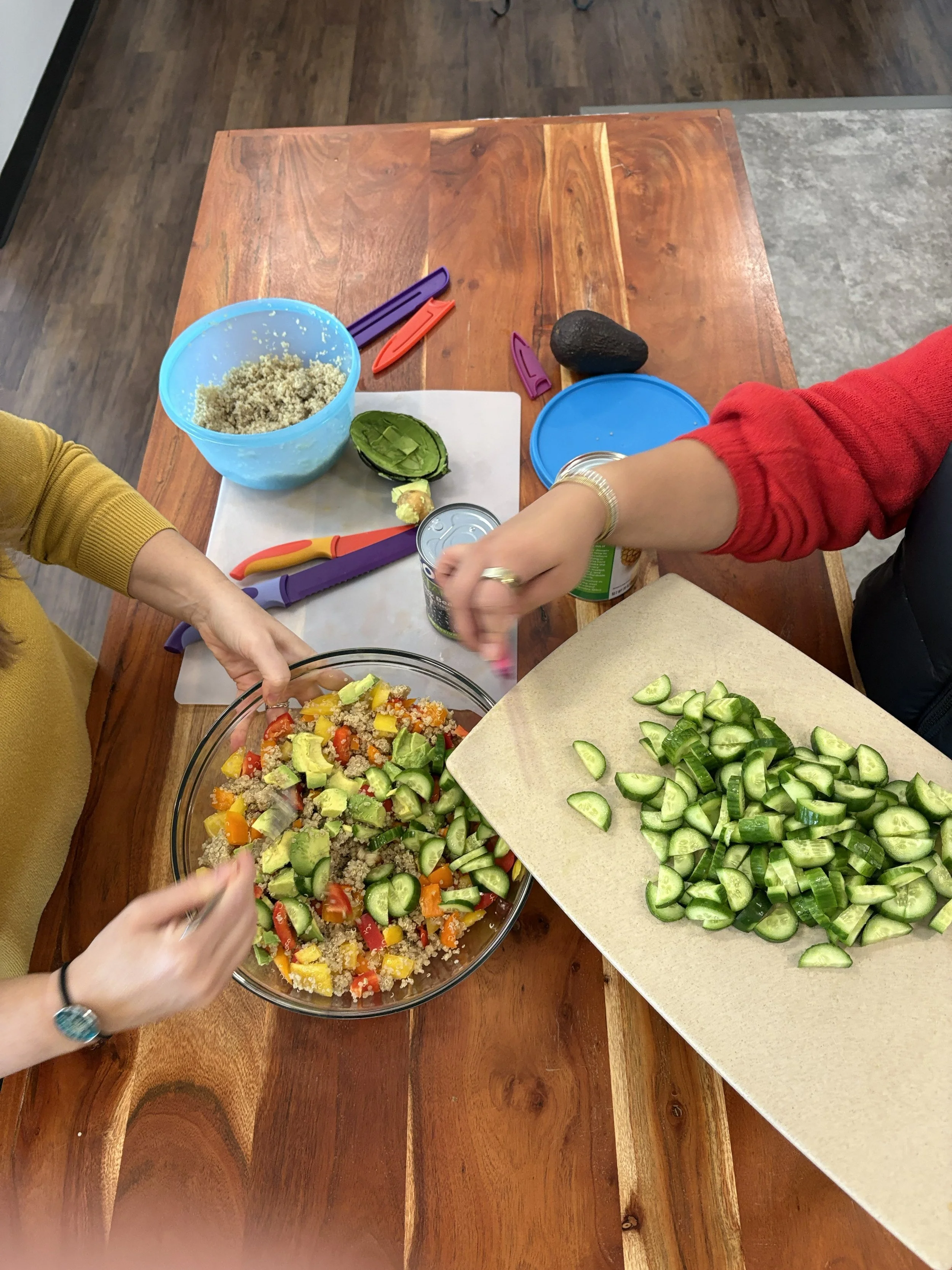 People preparing a vegetable salad with chopped cucumbers, tomatoes, yellow peppers, and cooked quinoa on a wooden table, with various tools and containers around.