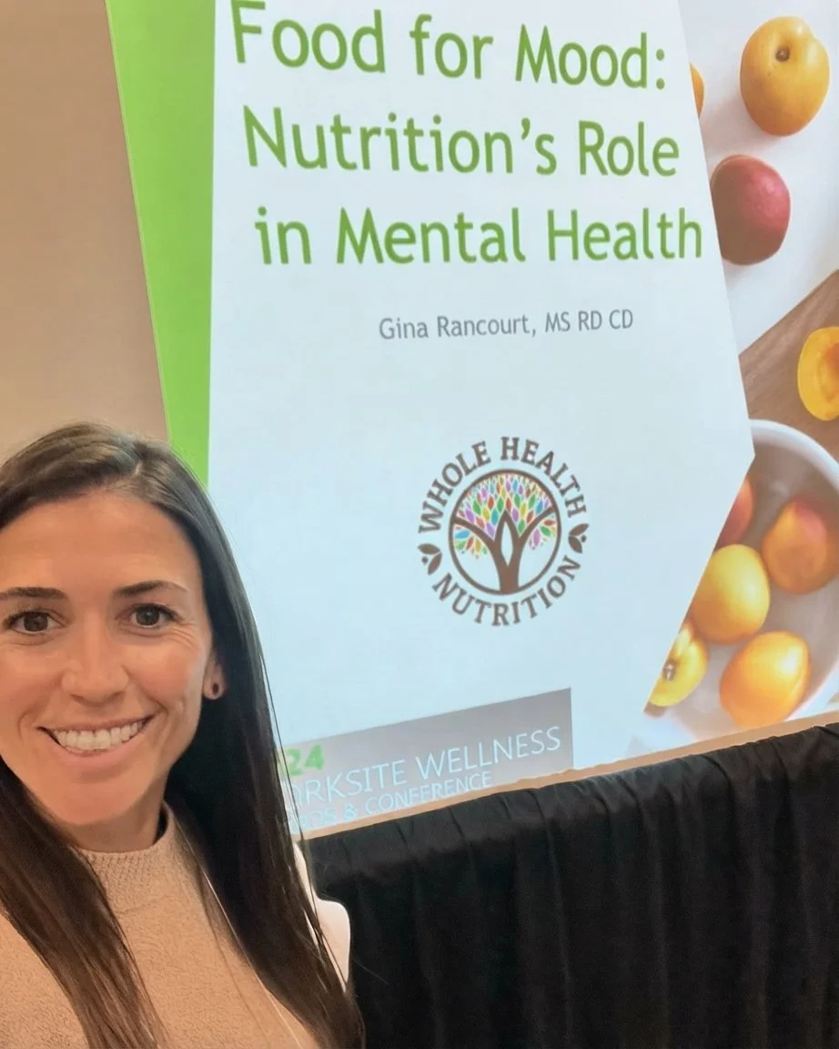 Smiling woman at a conference with a large presentation slide behind her. The slide displays the title 'Food for Mood: Nutrition's Role in Mental Health,'.