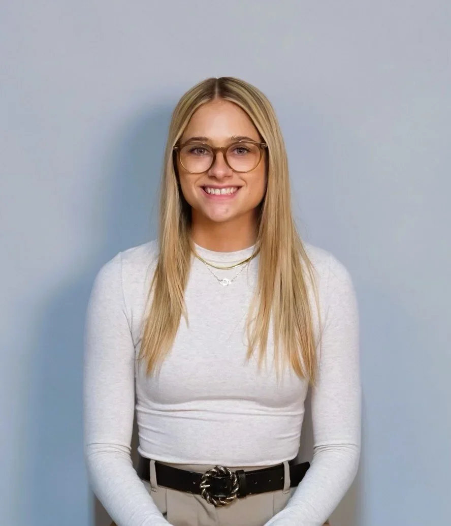 A young woman with long blonde hair, glasses, and a white long sleeve shirt standing against a plain light blue wall, smiling at the camera.