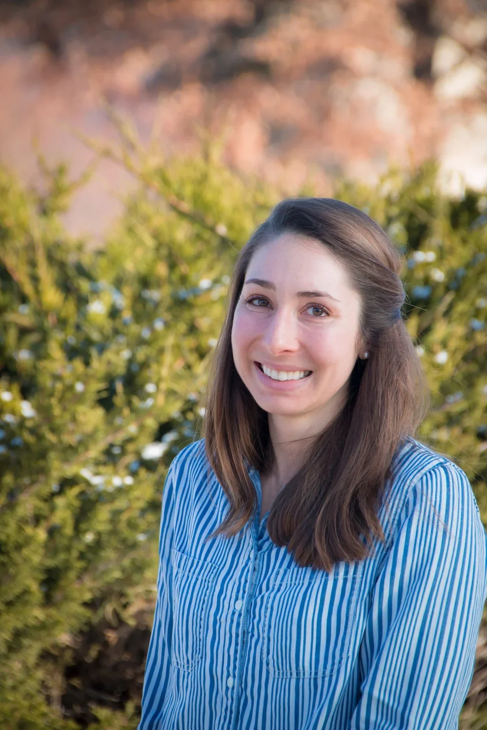 A young woman with brown hair and blue eyes smiling outdoors in front of green shrubbery and trees.