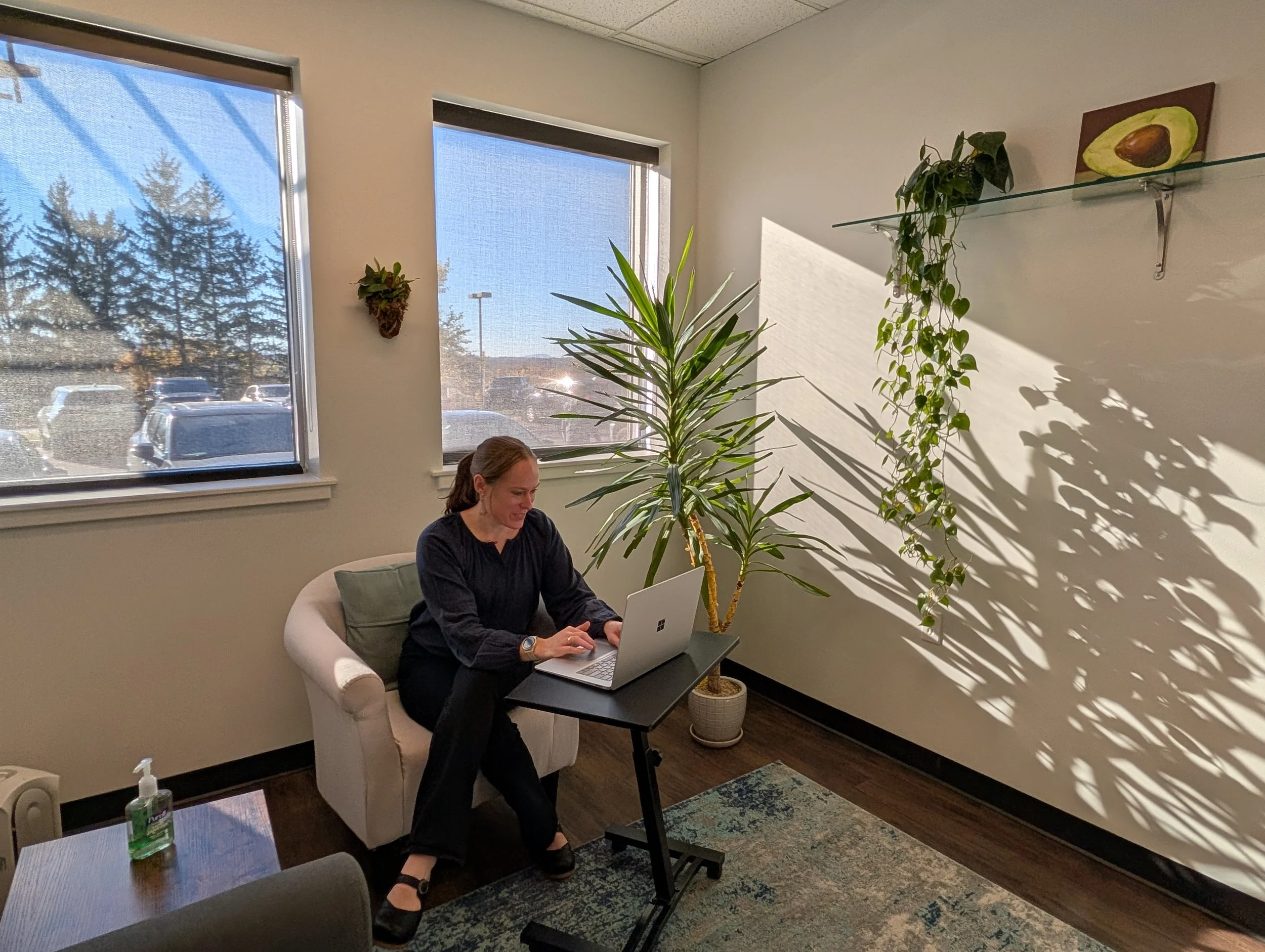 A woman sitting in a white armchair working on a laptop at a small black table in an office or waiting room. Behind her are two large windows with sunlight streaming in, casting shadows of plants and window frames on the wall. The room has light-colored walls, a potted plant, and artwork of an avocado seed on a glass shelf.