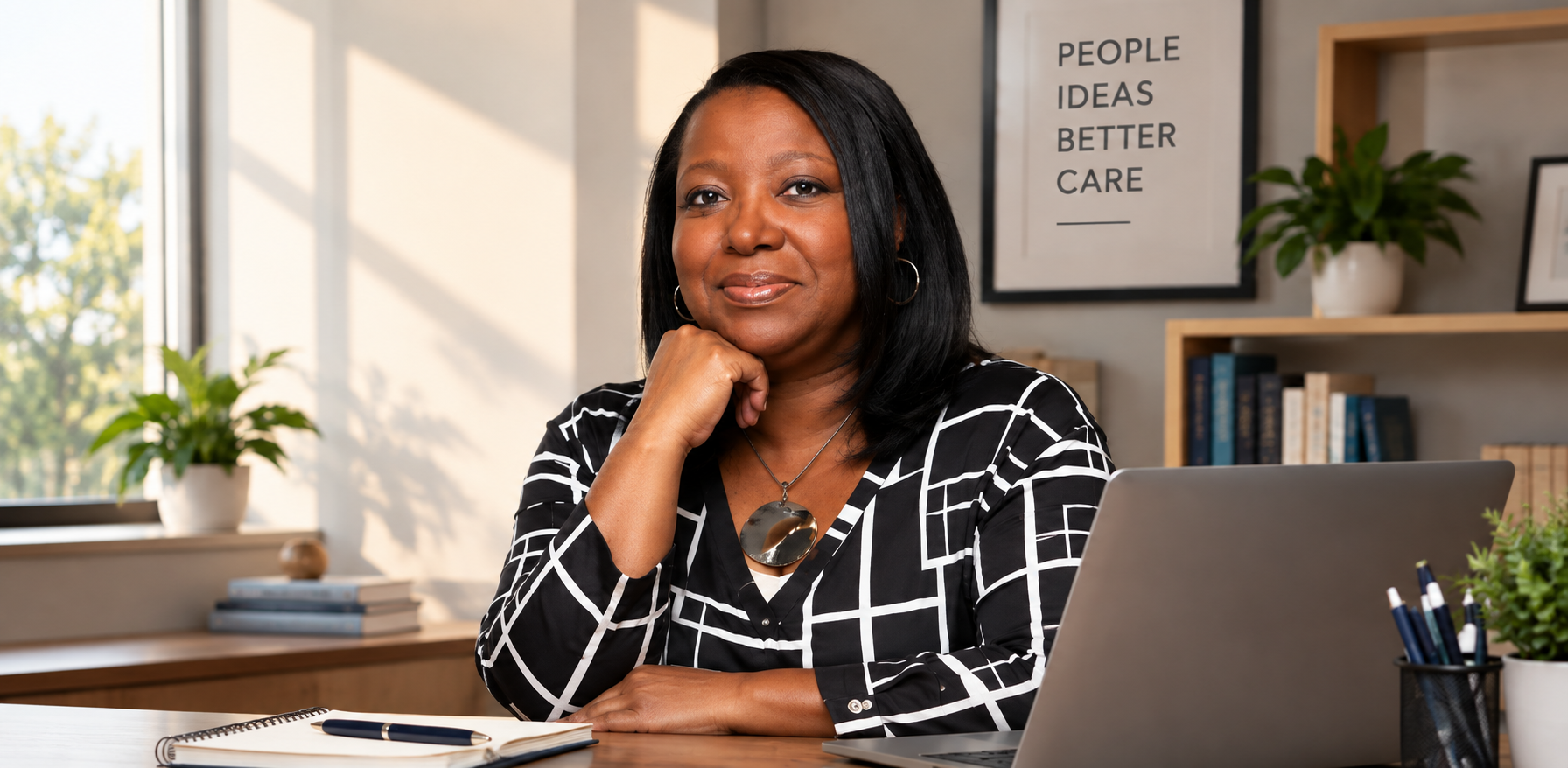 A woman sitting at a desk in an office, looking confidently at the camera with her hand resting on her chin. Behind her, there are bookshelves, plants, and a framed poster that reads 'People Ideas Better Care'.