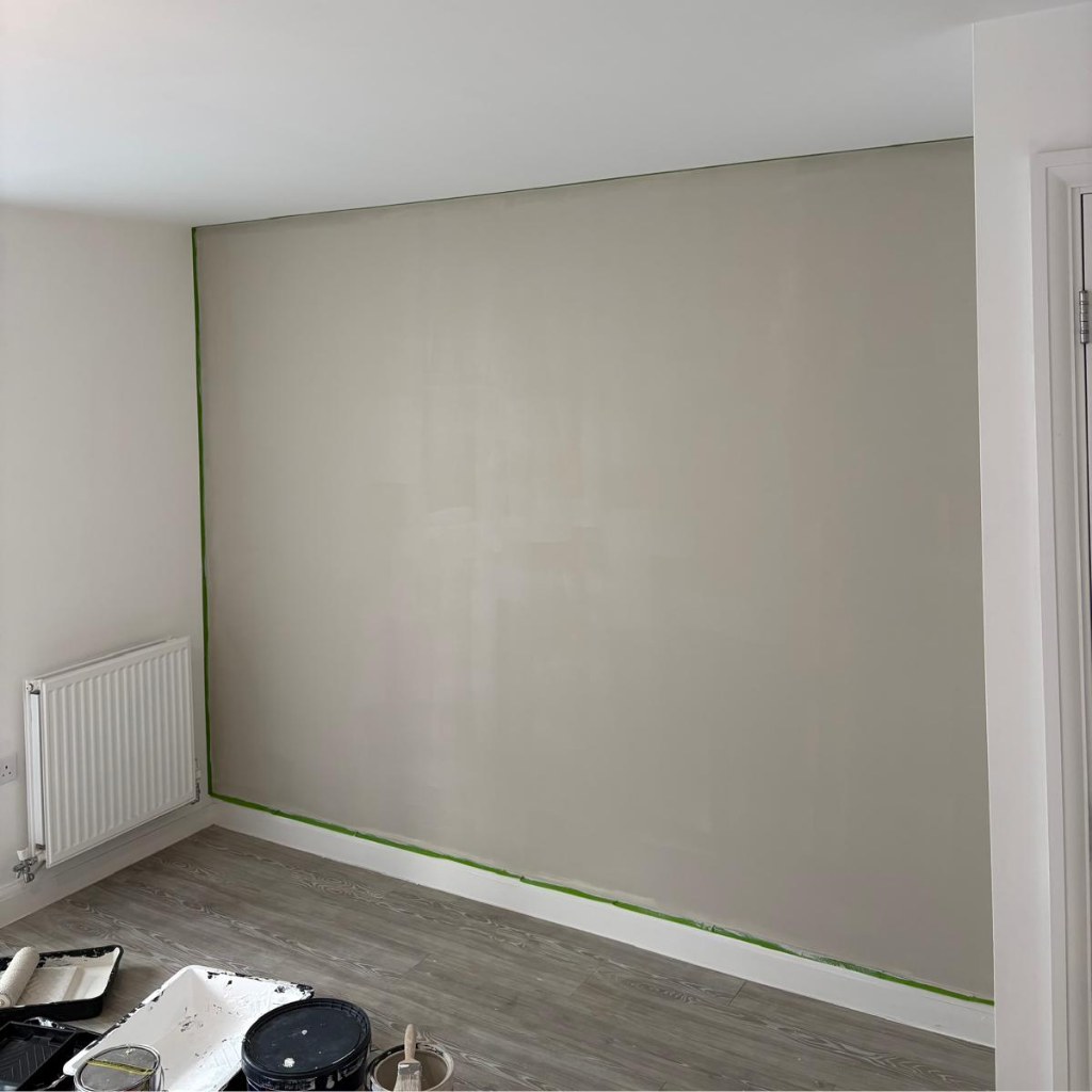 A room with a beige painted wall, painter's tape along the top edge, a radiator on the left, and painting supplies on the floor.