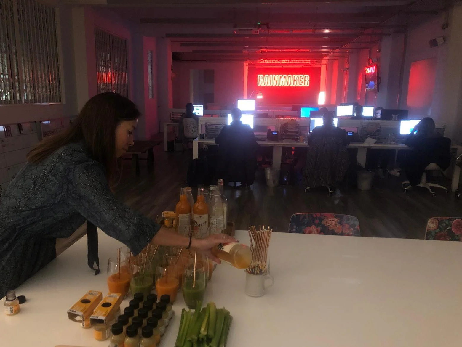 A woman pouring drinks at a counter with juice bottles, celery, and small boxes in an office setting with people working at computers in the background and a neon sign that says 'RADIOCRACKER'.