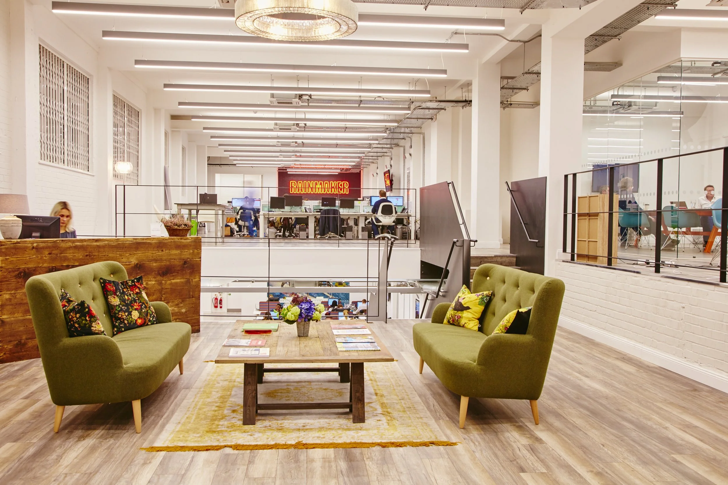 A modern office common area with seating, a wooden coffee table with magazines and colorful pillows, and a background with workspaces and a neon sign that reads 'Rainmaker.'