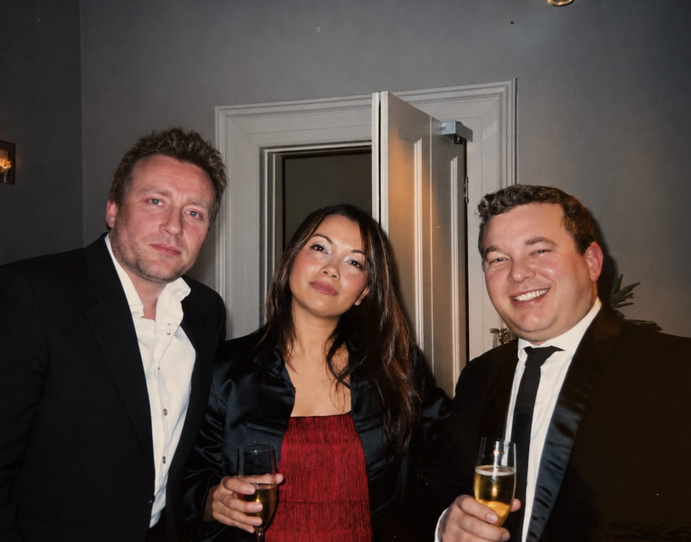 Three people dressed in formal attire at a social event, holding glasses of champagne and smiling at the camera, with a gray wall and open door in the background.