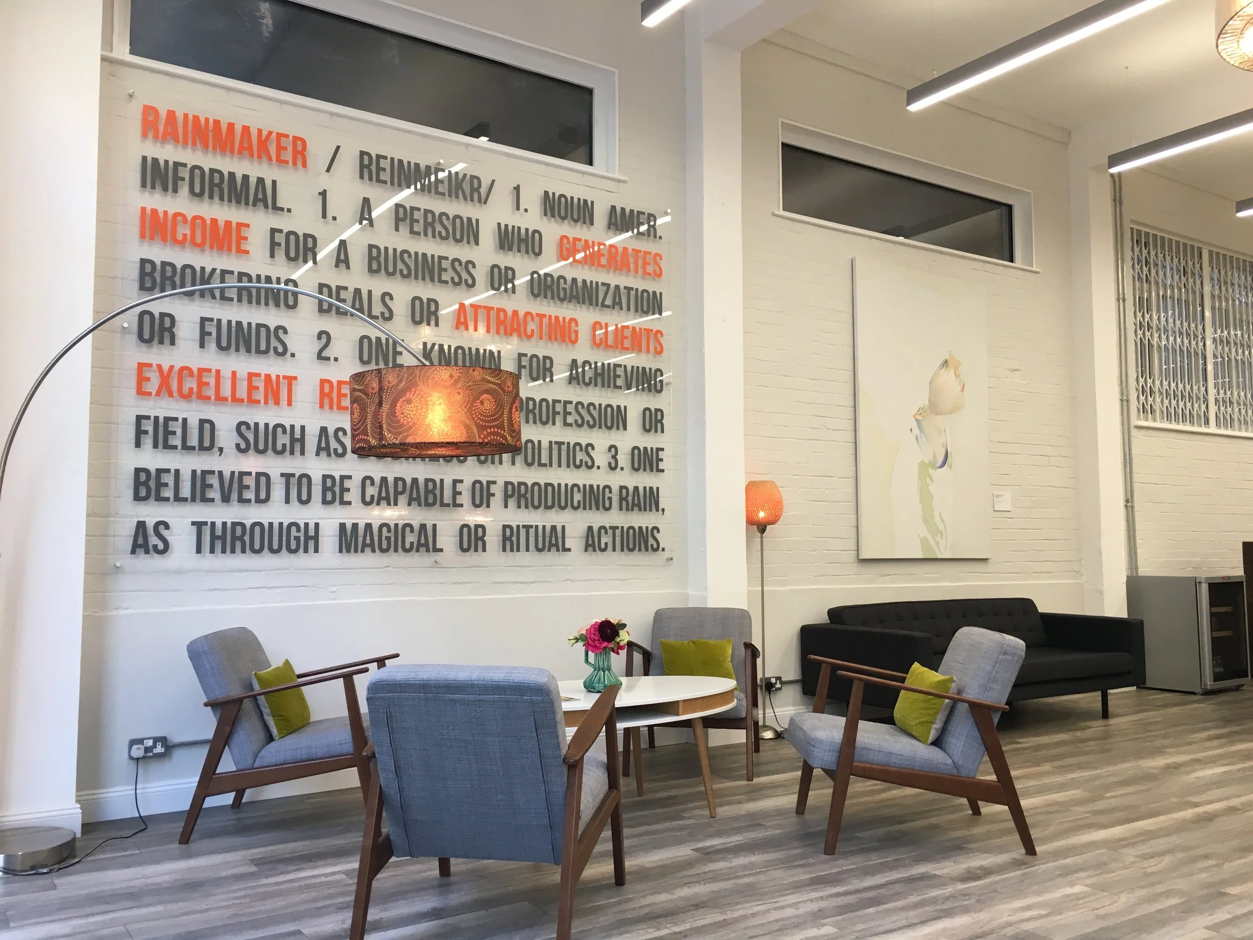 A modern, well-lit lounge area with multiple chairs, a small table with a flower arrangement, and a large wall display with text about rainmaking and related terms. There is a floor lamp, artwork, and large windows in the background.