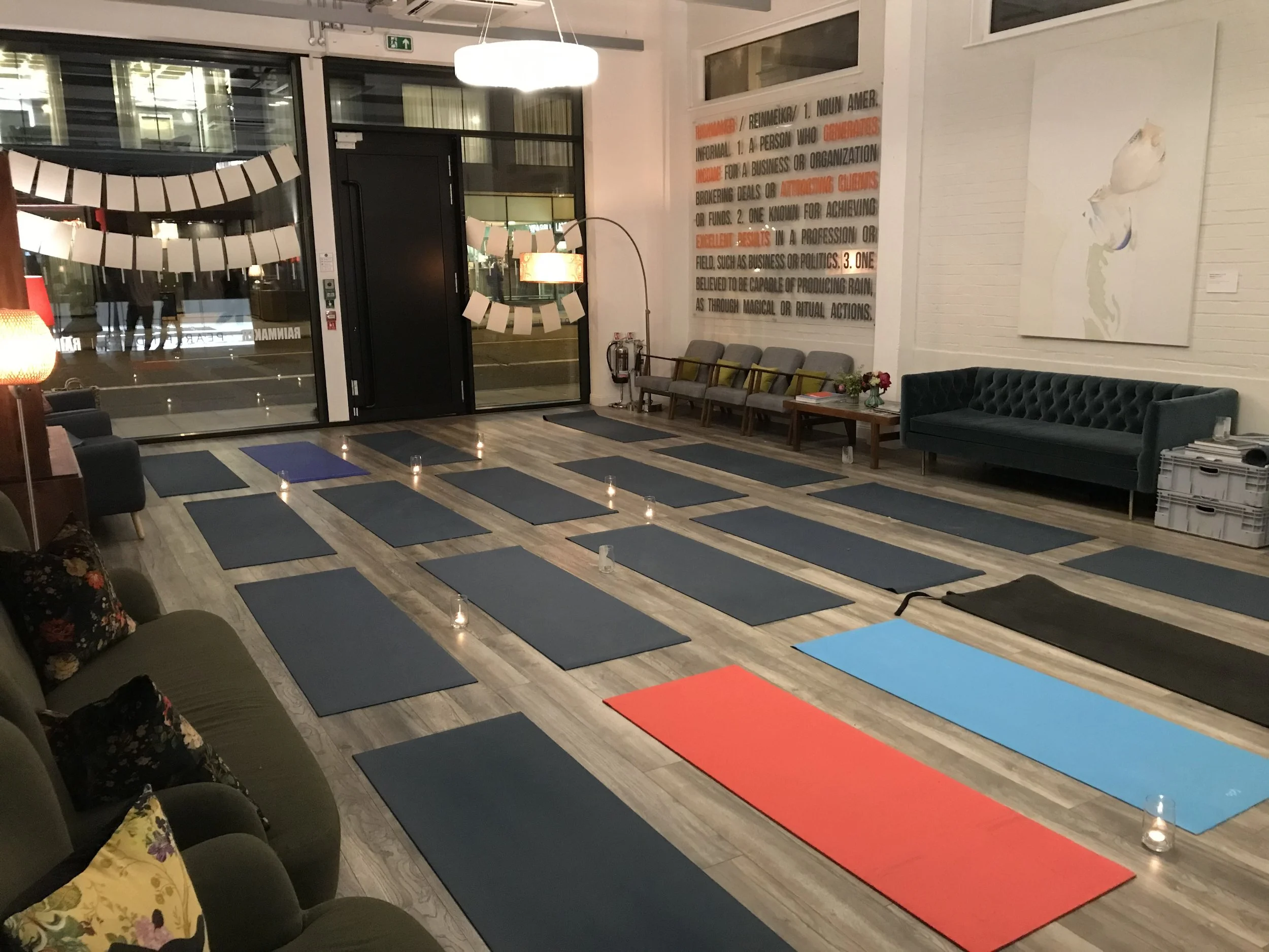 A yoga or meditation class setup in a cozy, well-lit space with yoga mats arranged on a wooden floor, candles along the edges, chairs along the wall for seating, a large white wall with colorful text, and artwork of a white horse painted on the wall.
