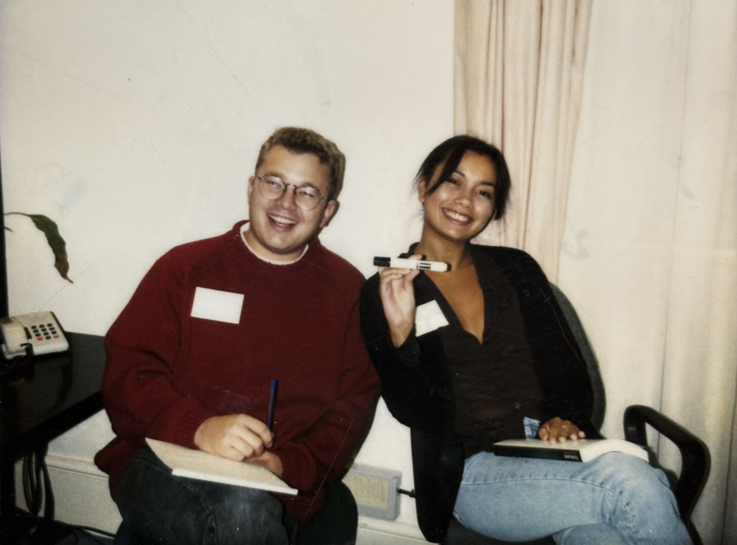 Two people, a man and a woman, sitting at a table in an indoor setting. The man has short brown hair, glasses, and is wearing a red sweater and holding a marker. The woman has long dark hair, visible earrings, and is wearing a black jacket. She is ho