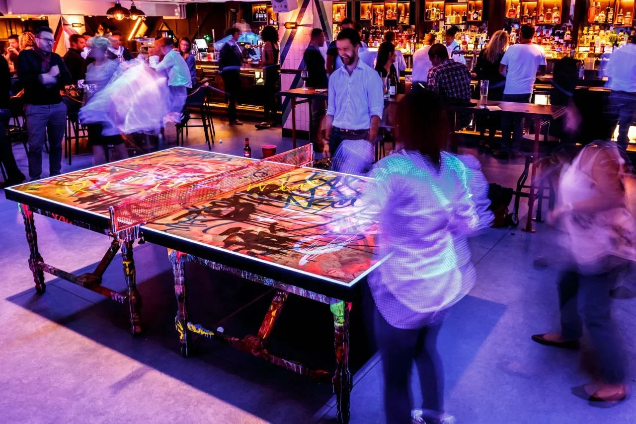 People playing ping pong in a vibrant, lively bar with dim, colorful lighting and a busy bar in the background.