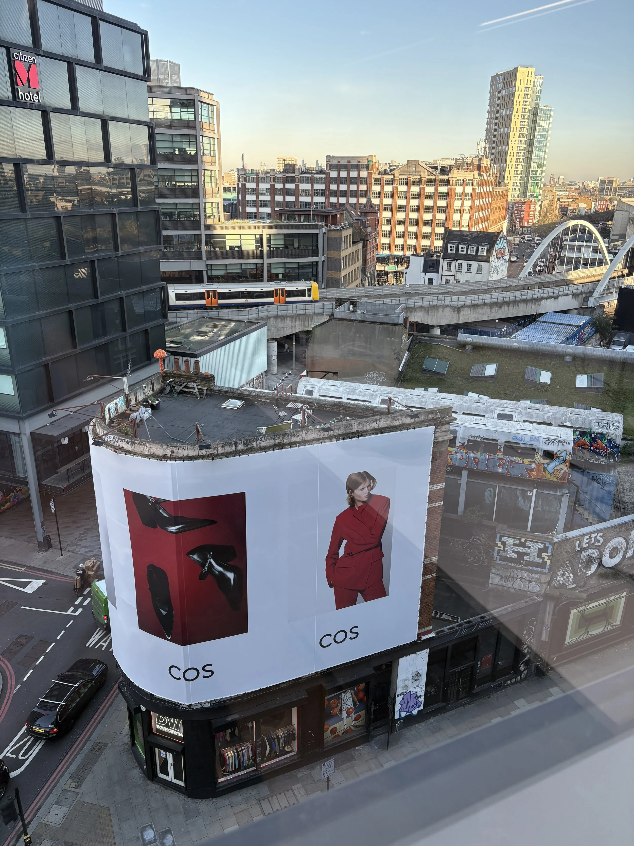 Cityscape view from a high floor, showing modern buildings, a tram on an elevated track, and large billboards including one for COS featuring a woman in a red suit.