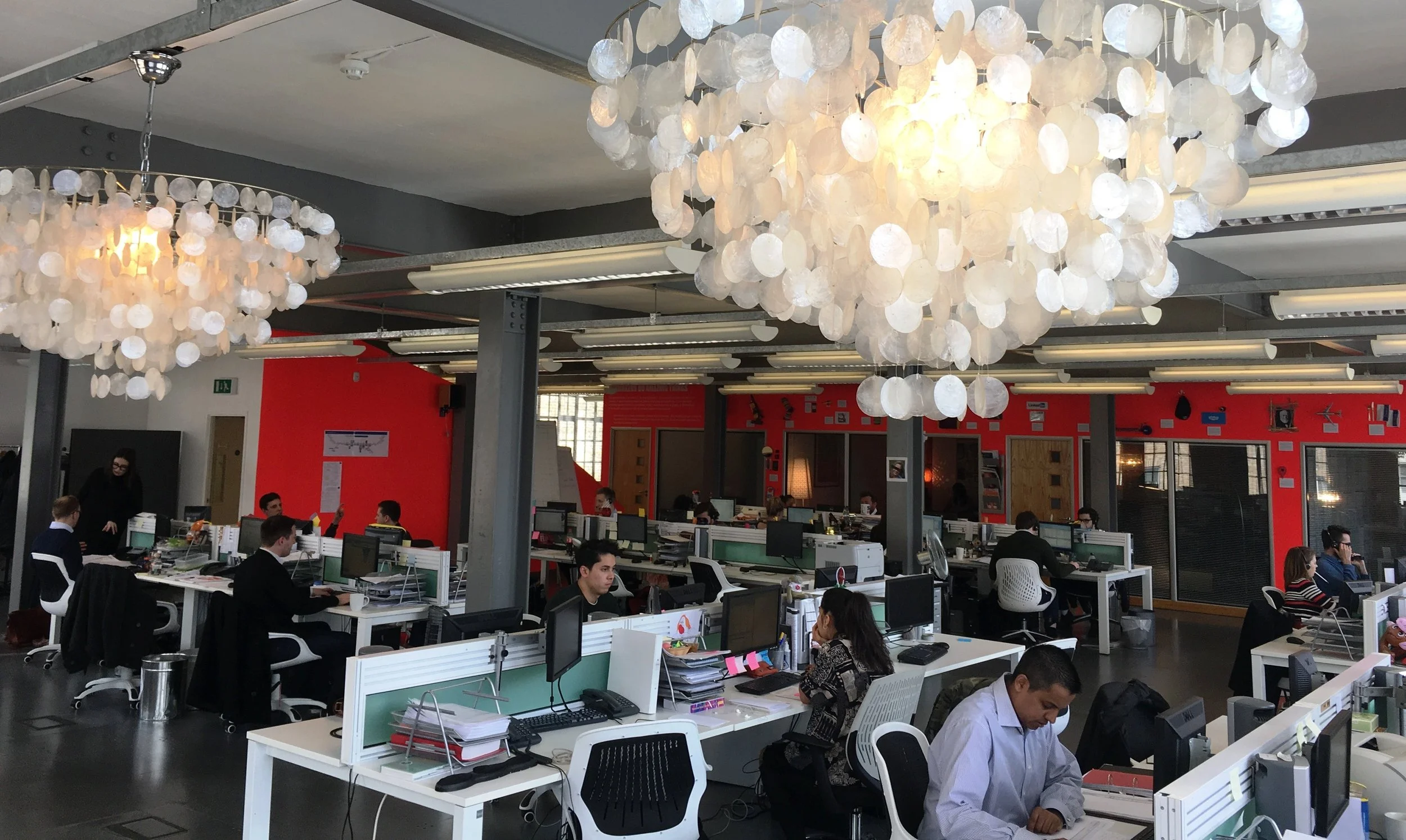 Modern open-plan office with workers at desks, multiple computer monitors, and large decorative white chandeliers hanging from the ceiling.